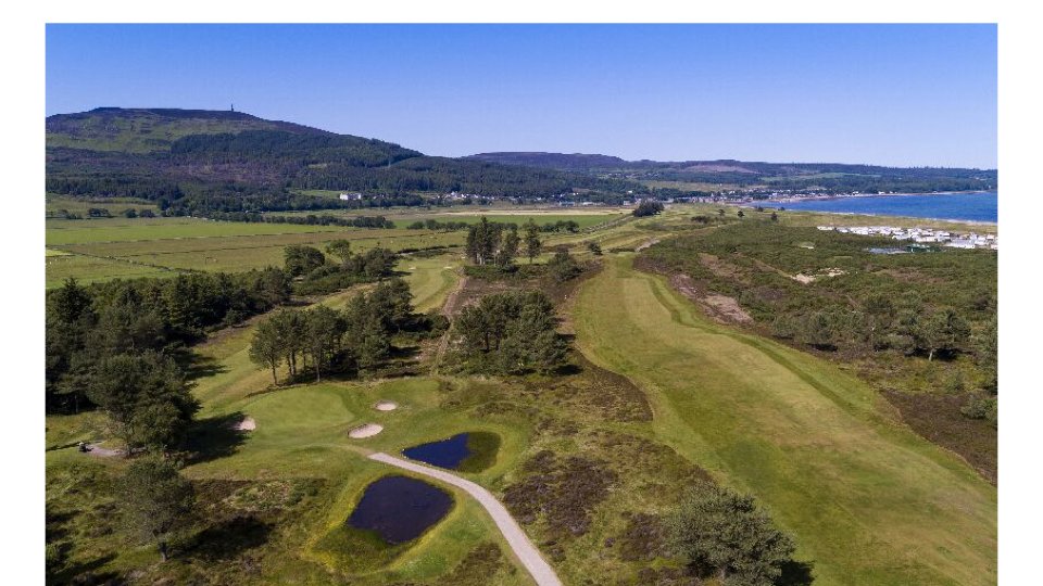 Aerial view of a scenic golf course with sand traps, water hazards, lush green fairways, trees, rolling terrain, mountains, coastline, and residential buildings in the distance.