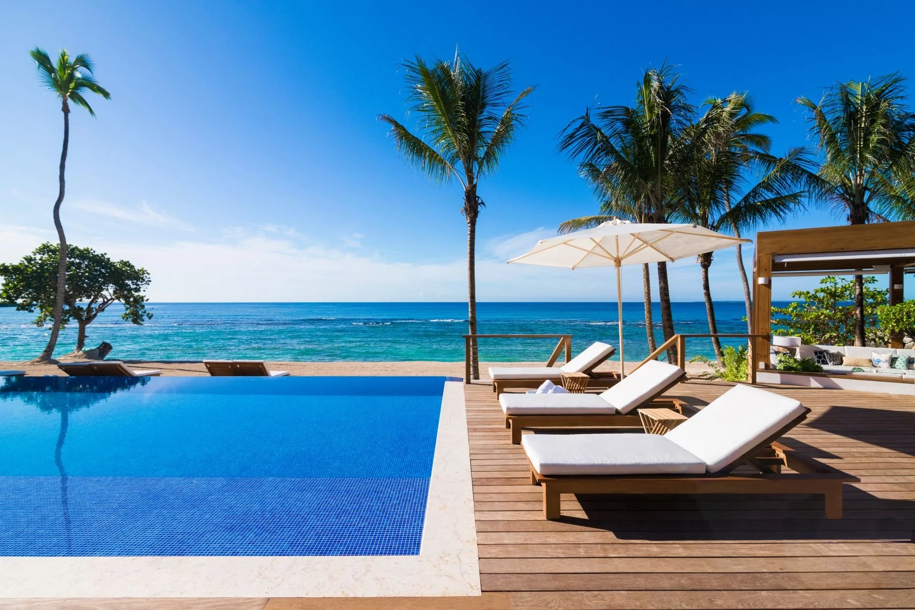 Beachside pool area with wooden deck, white lounge chairs under umbrellas, and palm trees overlooking the ocean.