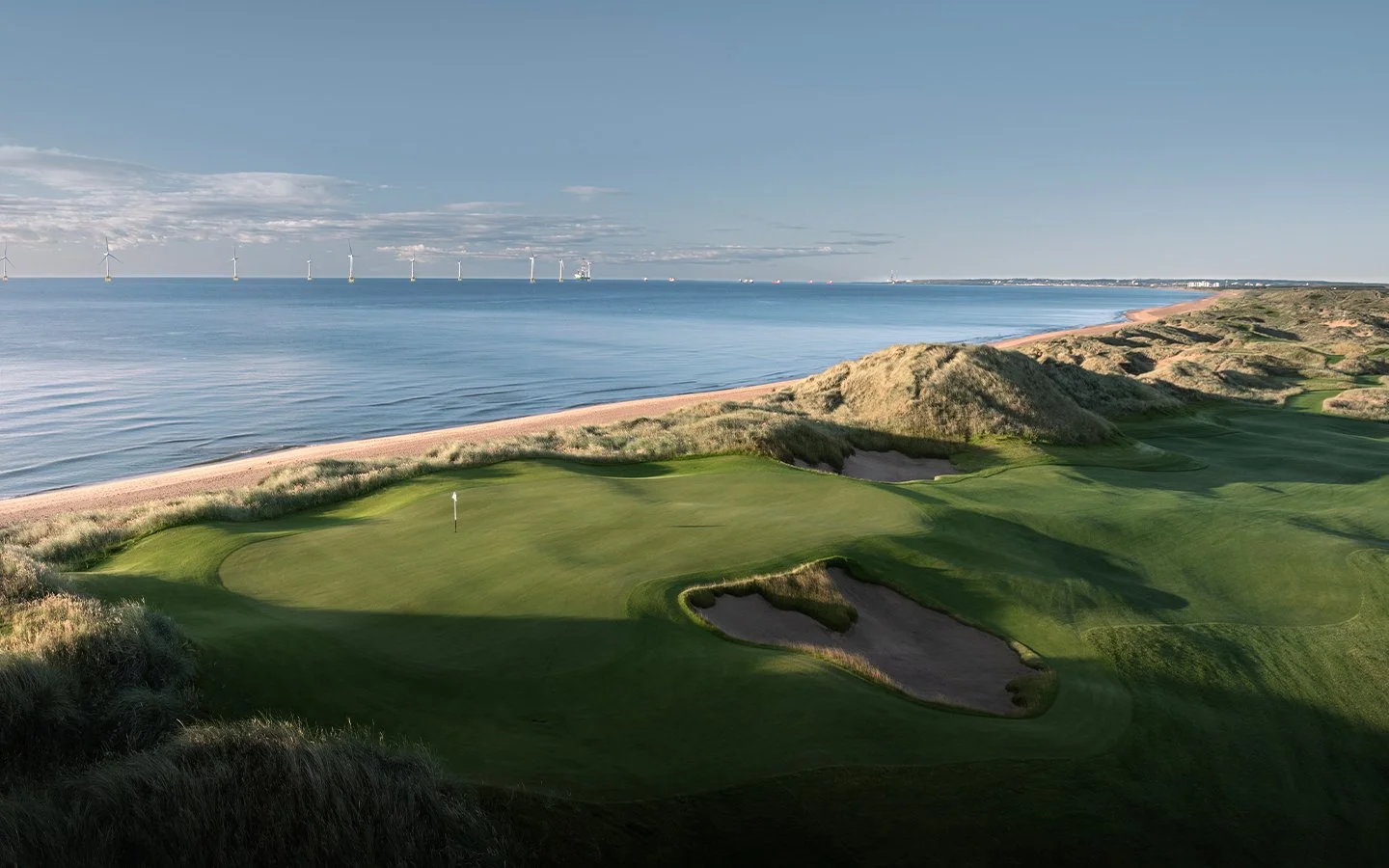A golf course on the coast with a green, sand traps, and dunes, overlooking the ocean with wind turbines in the distance under a partly cloudy sky.