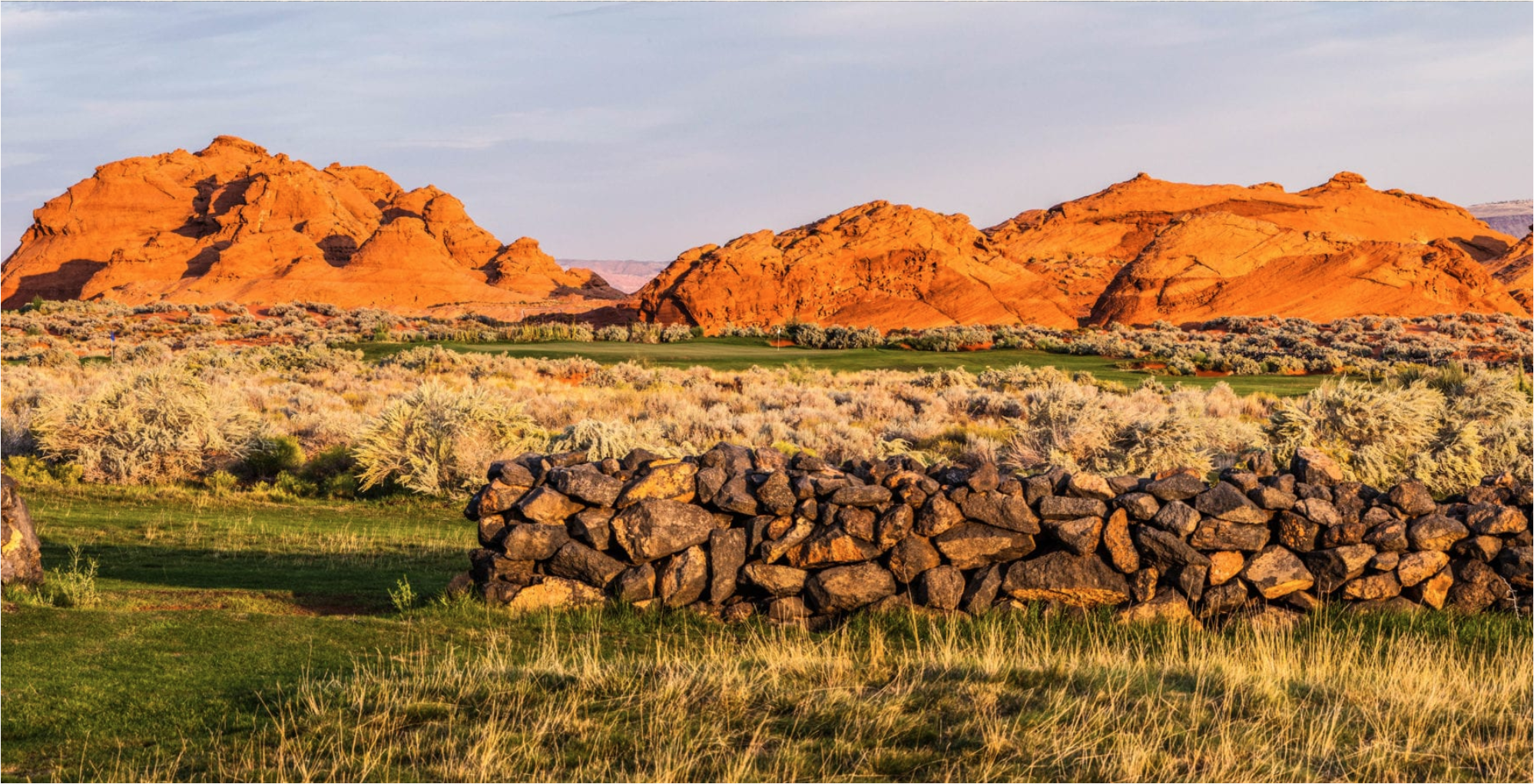 A desert landscape with large orange rock formations in the background, a stone wall in the foreground, and sparse desert vegetation.