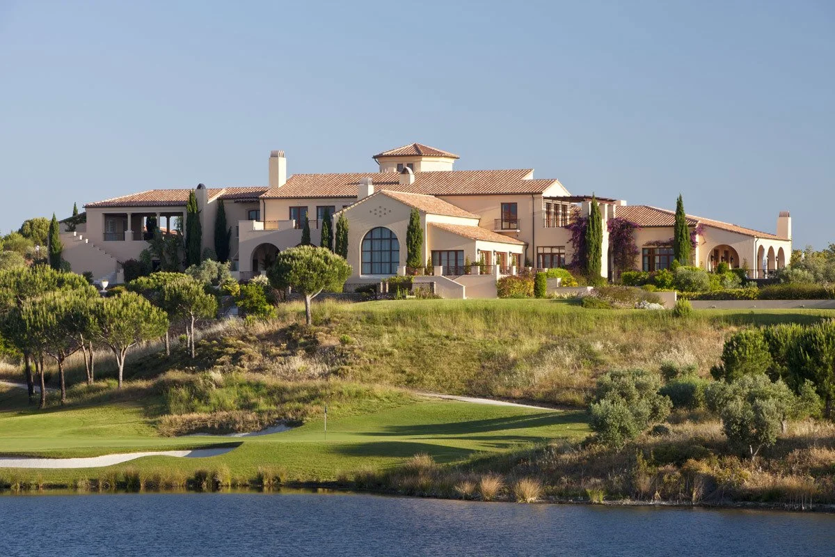 Luxurious estate with a Mediterranean-style house, surrounded by trees and landscaping, overlooking a golf course and a body of water under a clear blue sky.