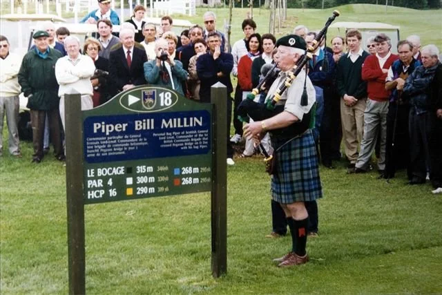 A man in a kilt playing bagpipes in front of a group of people gathered around a sign at a golf course. The sign reads 'Piper Bill Millin' and provides details about his connections to D-Day and his military service.