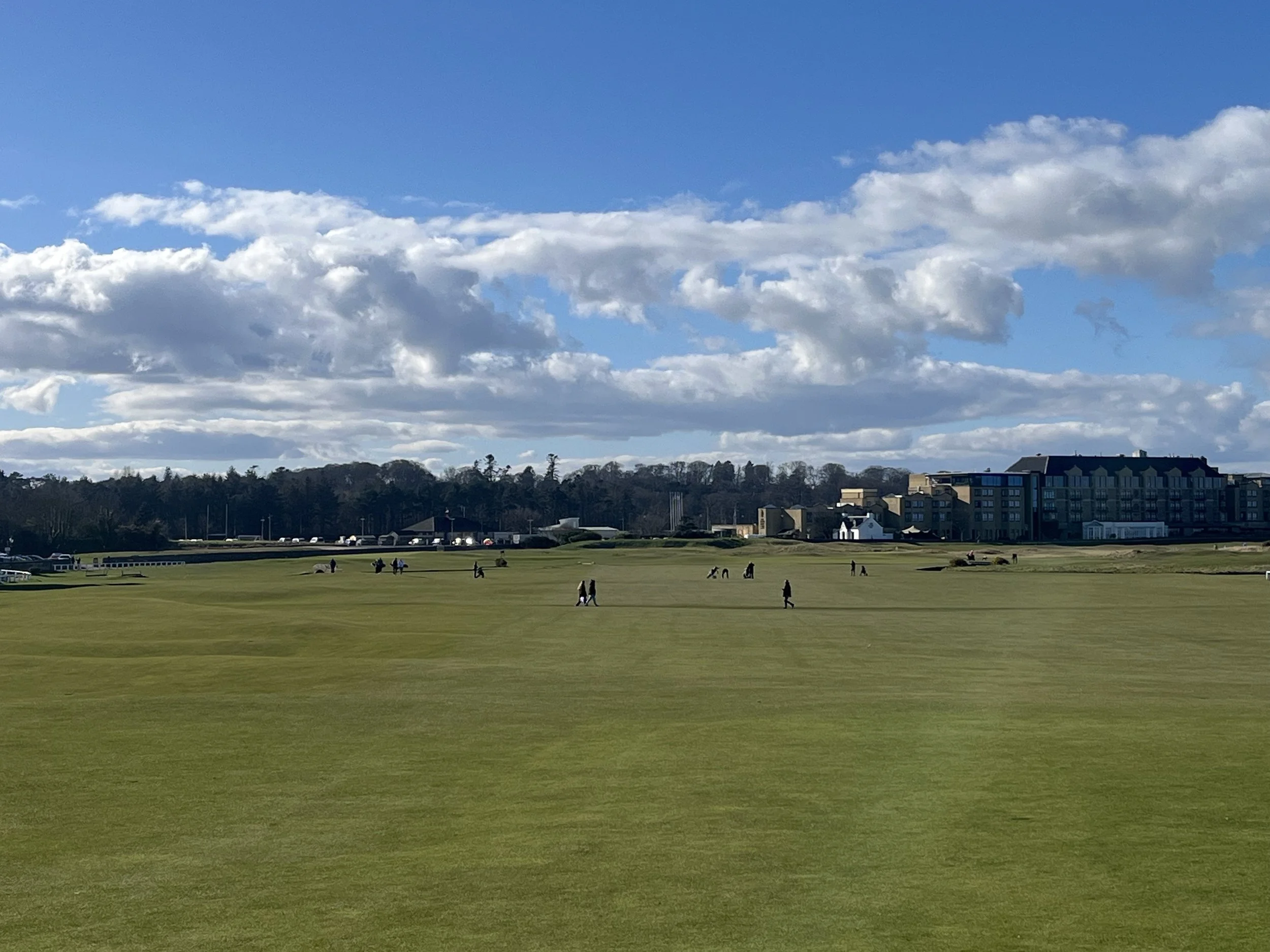 A scenic view of golfers playing  the iconic first hole (The Burn) at the Old Course, St Andrews, Scotland, on a beautiful sunny day. The wide green fairway stretches toward the prominent Old Course Hotel in the background under blue skies.