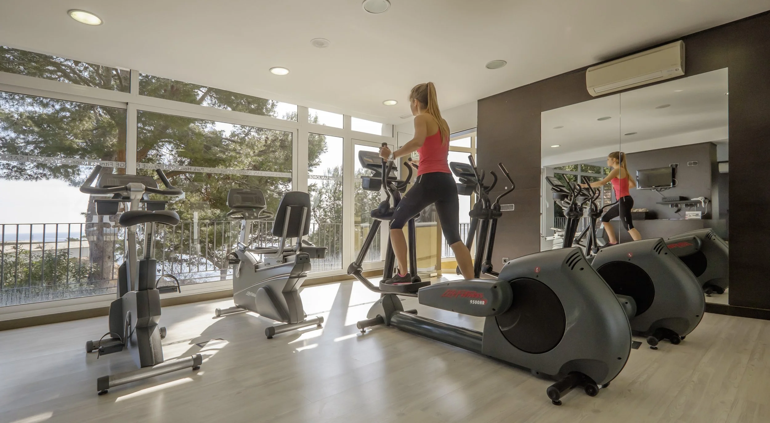 Woman using an elliptical machine in a bright, modern gym with large windows showing trees outside. Mirrors reflect her and the equipment.