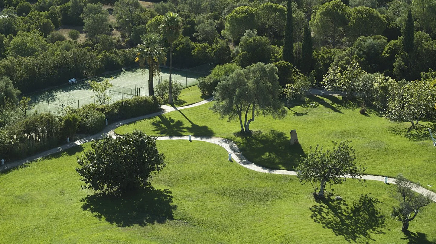 Aerial view of a park with a winding pathway, multiple trees, grassy areas, and a tennis court in the background.