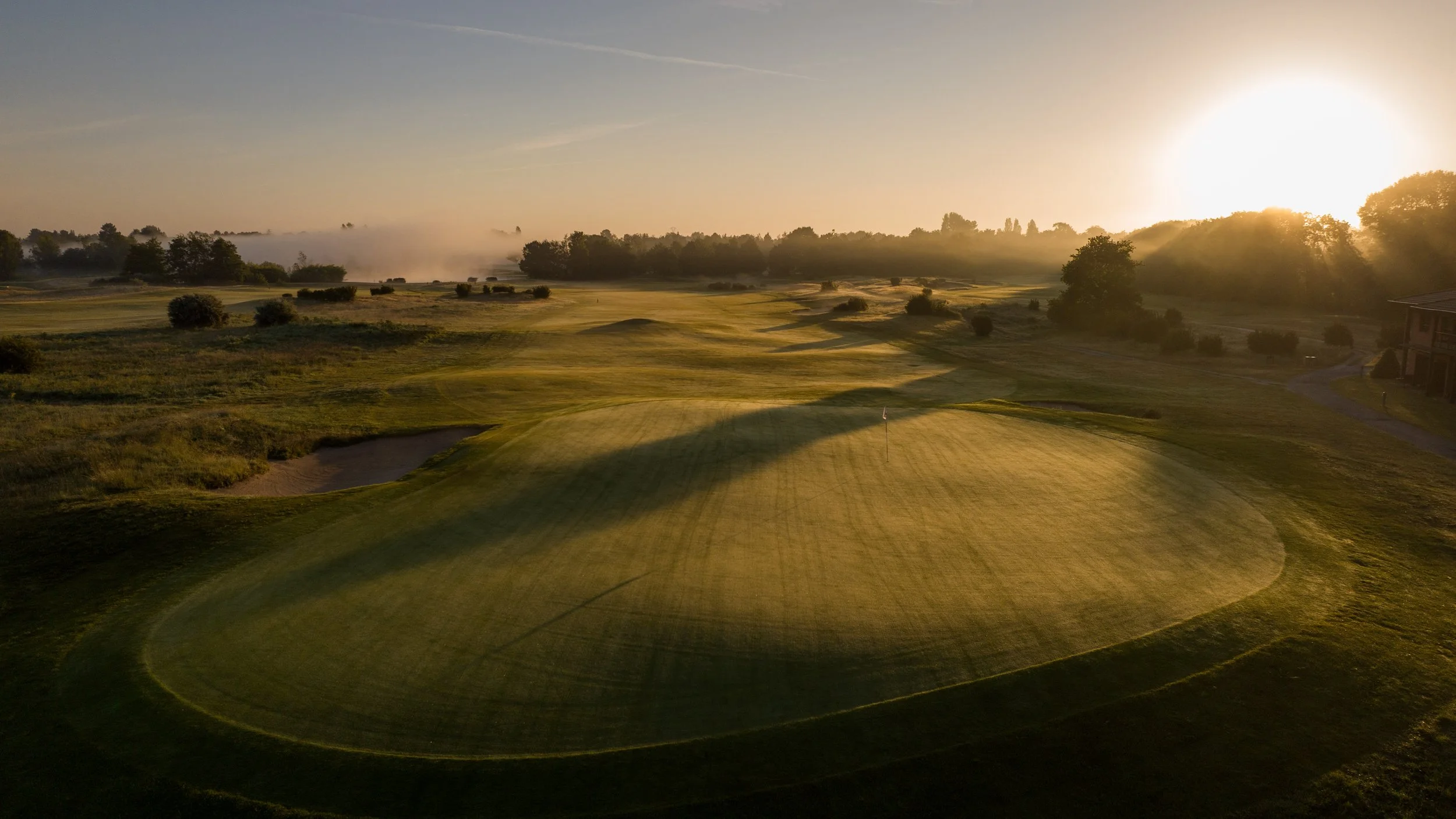A golf course at sunrise with a green, sand trap, and trees in the distance.