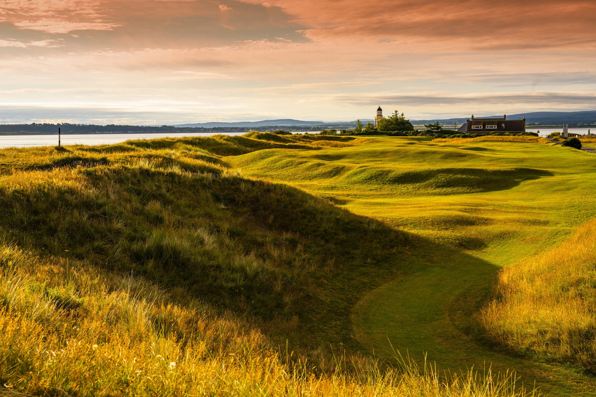 Scenic view of a grassy landscape with rolling hills, a lighthouse, and a house near the water during sunset, with a cloudy sky and distant mountains.