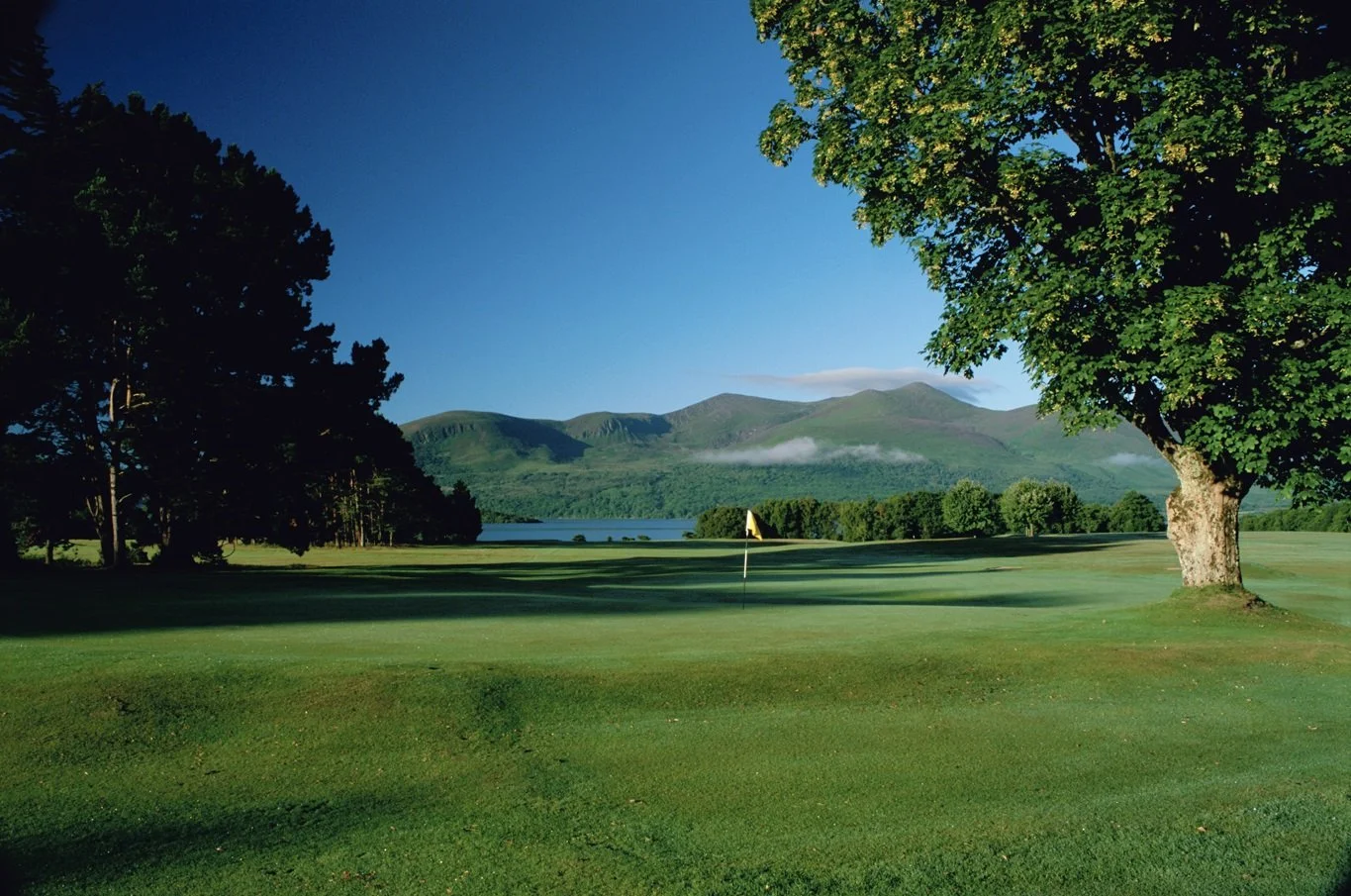 A golf course with a green and water in the background, surrounded by trees and mountains under a blue sky.