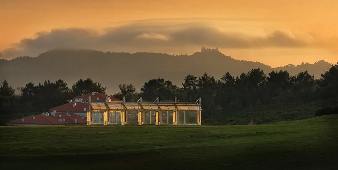 A glass-enclosed structure in a grassy area with a house nearby, with a mountain and a castle silhouette in the background at sunset.