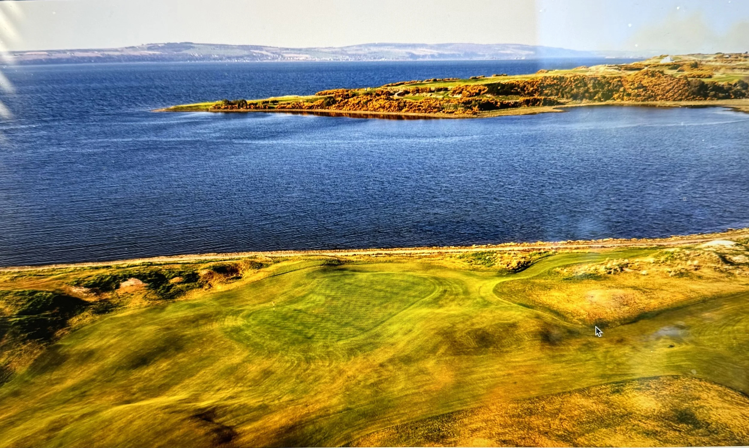 A scenic aerial view of a lake with bright blue water, surrounded by green and brown land, possibly a golf course, with hills or cliffs in the distance.