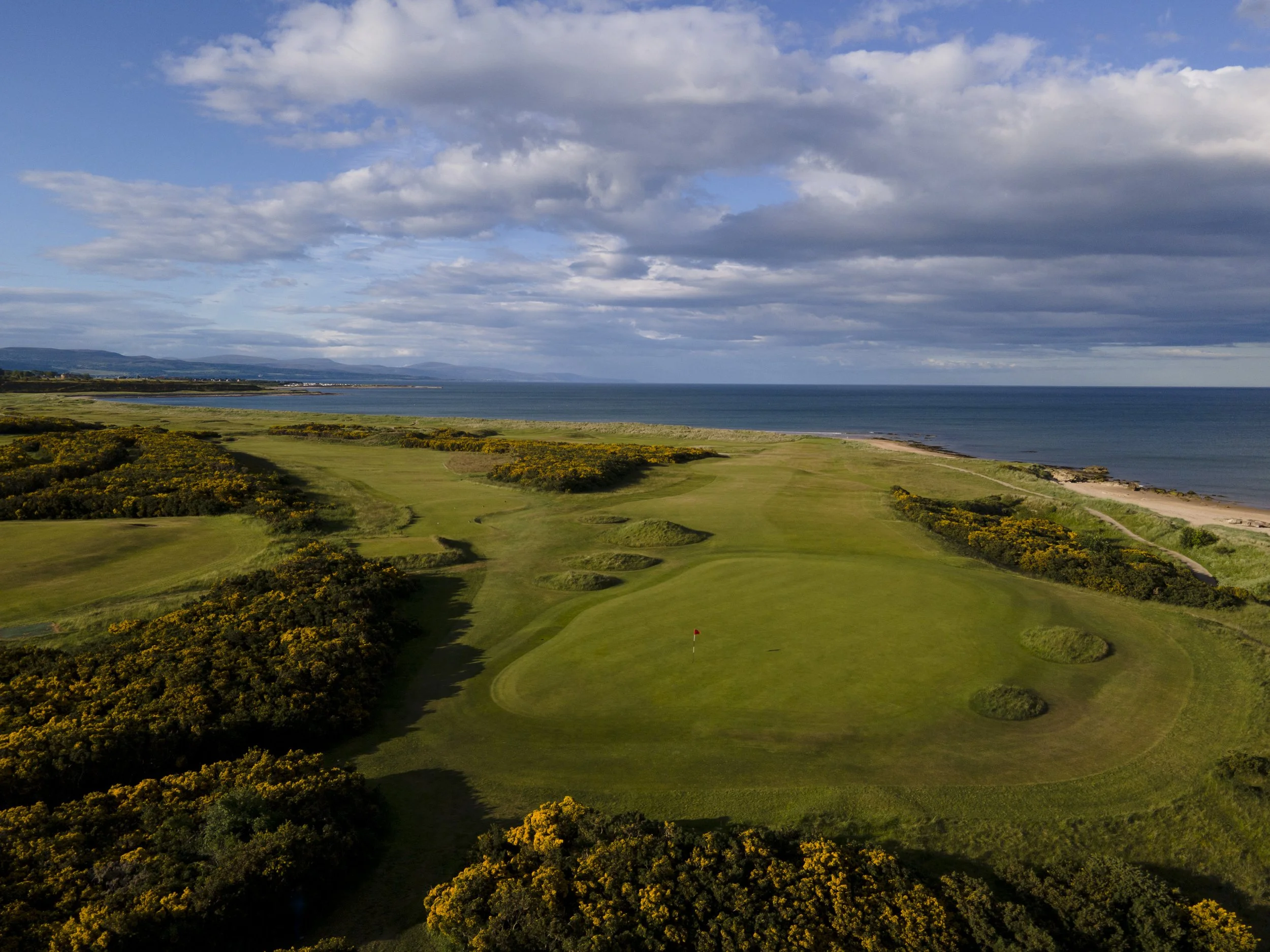A coastal golf course with green fairways, sand traps, and a flag, overlooking a body of water and land in the distance under a partly cloudy sky.