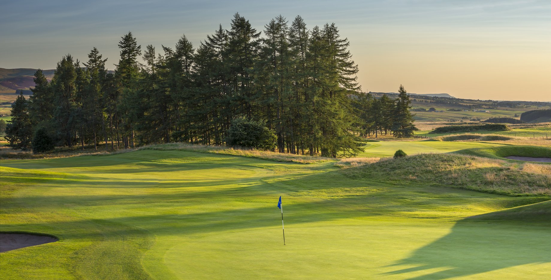 A golf course with green grass, a flag on the putting green, trees in the background, and a sunset sky.