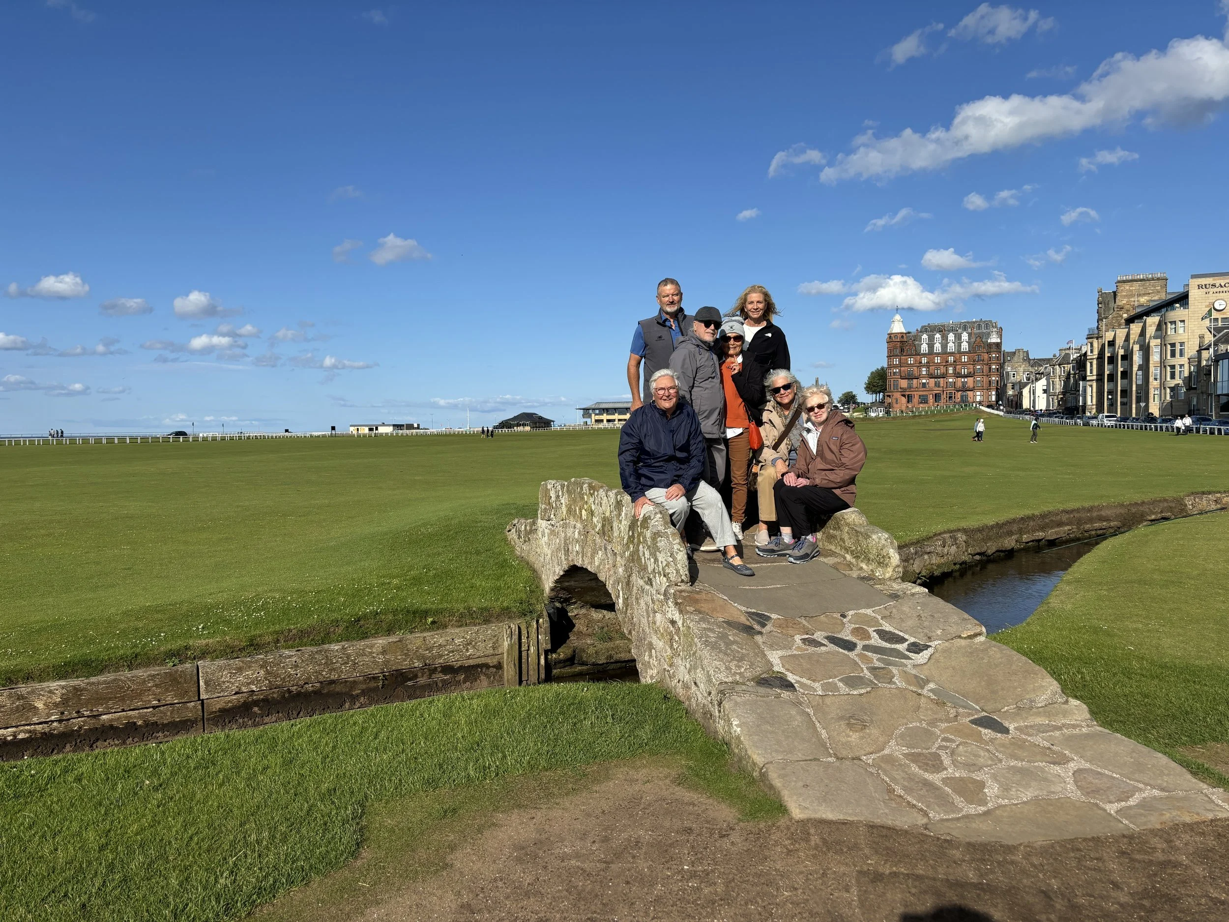 Group of eight seniors on a small stone bridge over a stream at a grassy park, with historic buildings and a blue sky in the background.