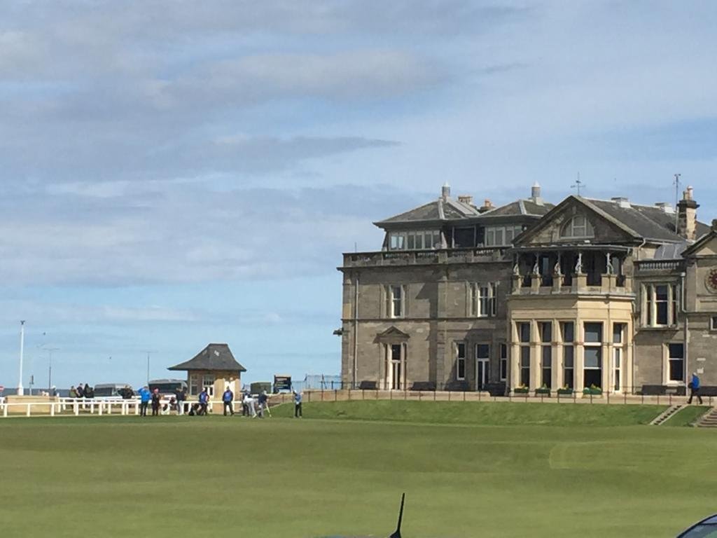 A large historic stone mansion with multiple stories and elaborate architecture, located on a well-manicured golf course with a group of people nearby, under a partly cloudy sky.