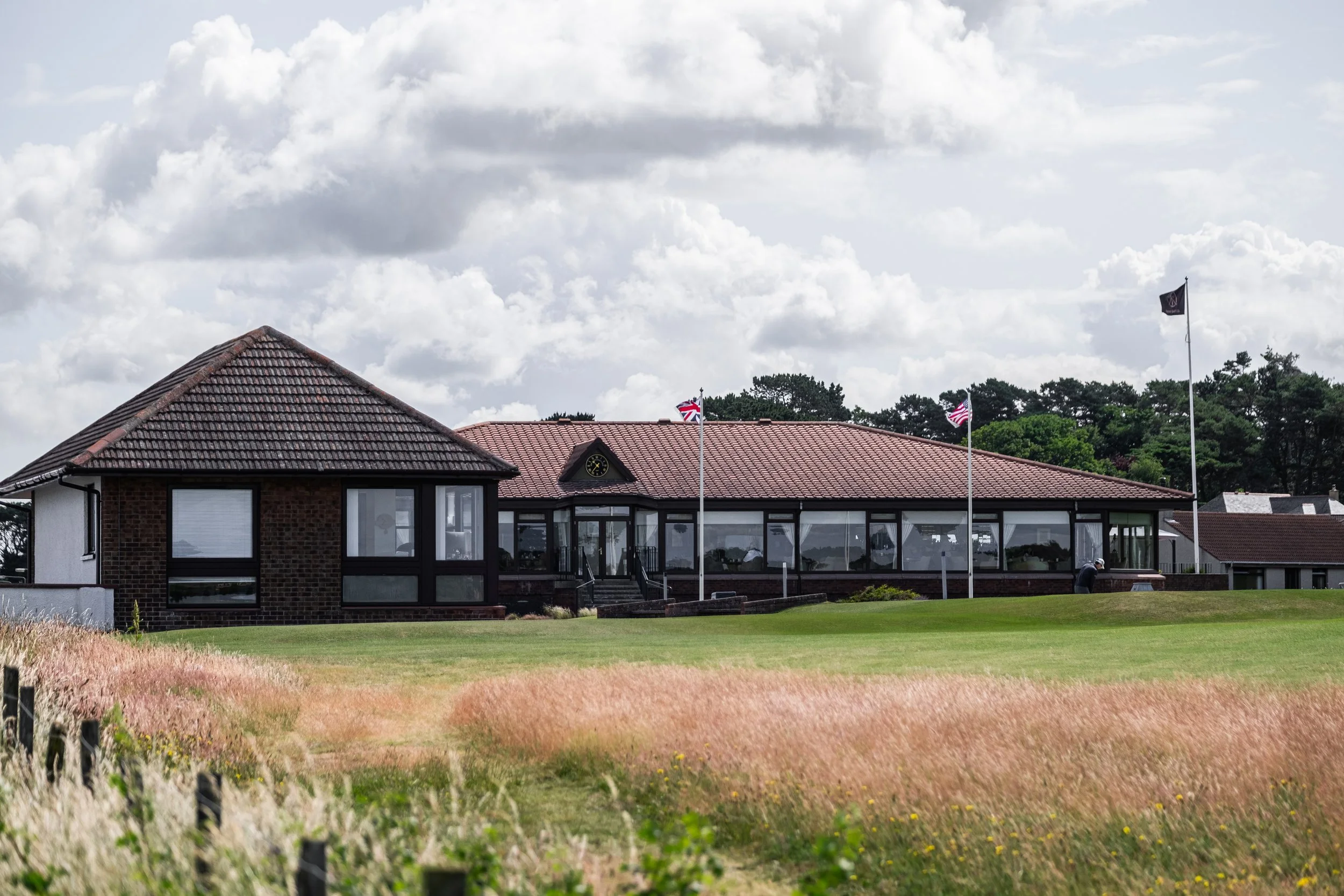 A building with a tiled roof, large windows, and a clock, surrounded by grass and trees, with flags in the foreground and a cloudy sky above.