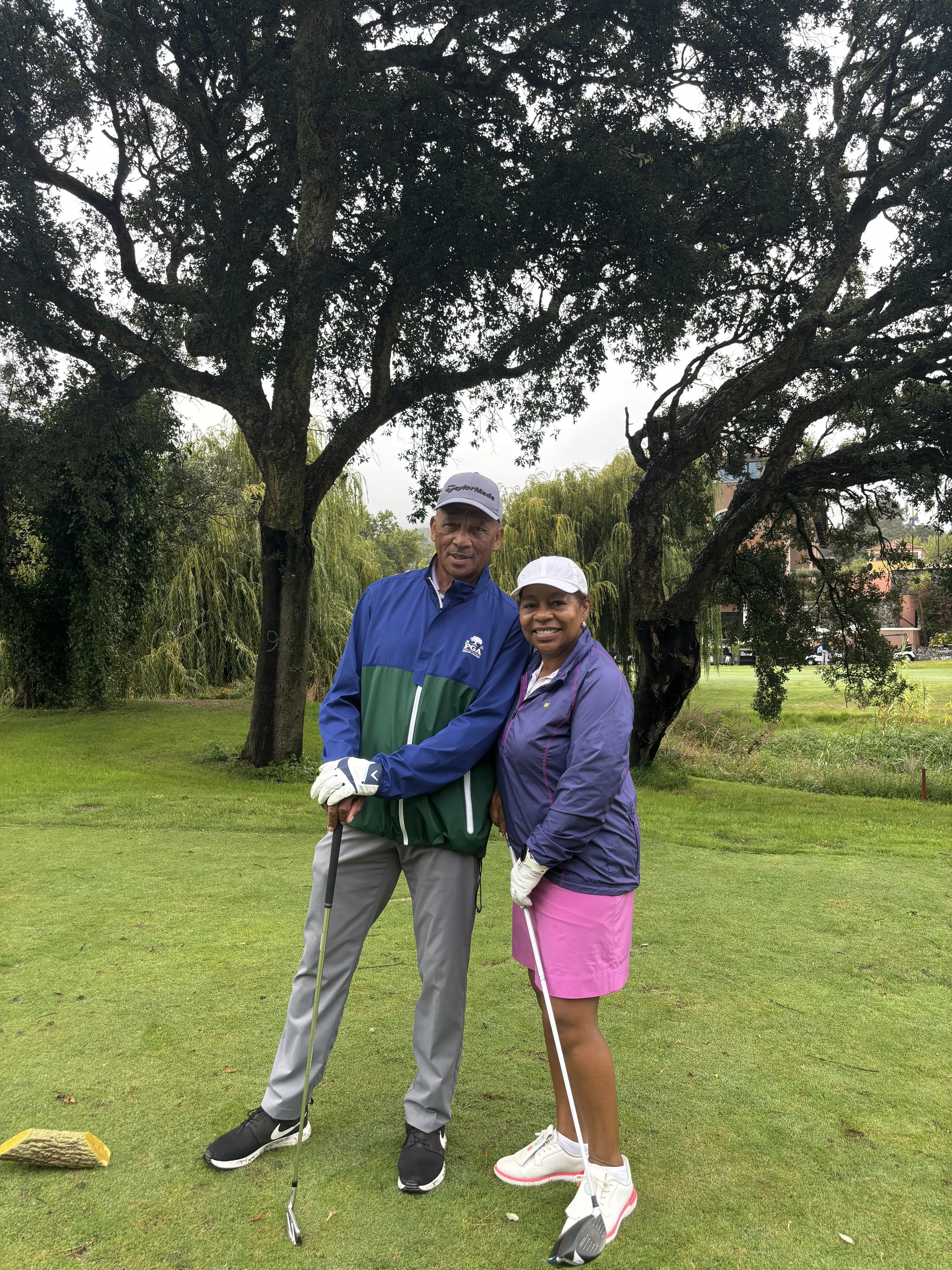 A man and woman standing on a golf course holding golf clubs, smiling, with trees and a grassy area in the background on a cloudy day.