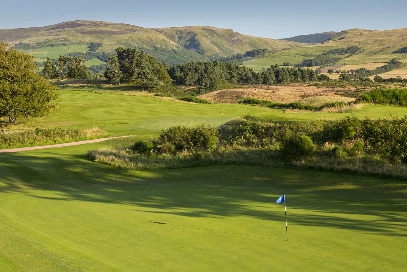 A lush green golf course with a blue flag on the putting green, surrounded by trees, shrubs, and rolling hills in the background under a clear sky.