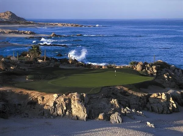 A golf course with a green near the rocky coastline and ocean waves in the background.
