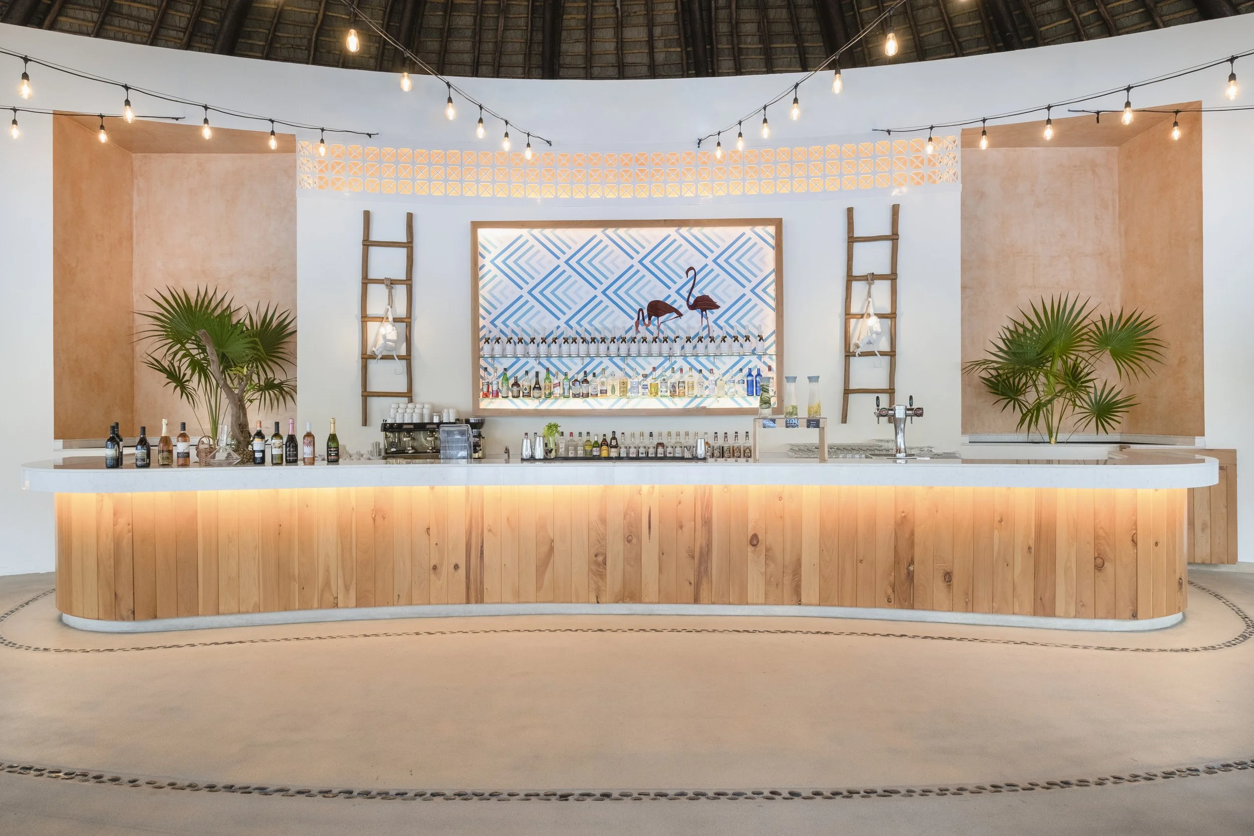 Beach bar with wooden counter, tropical plants, string lights, and a colorful geometric background featuring flamingos and a bar area with bottles and glasses.