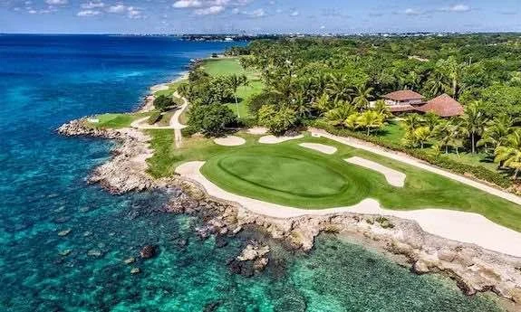 A golf course located on a rocky coastline, overlooking clear blue ocean water, with trees and a building with a red roof nearby.