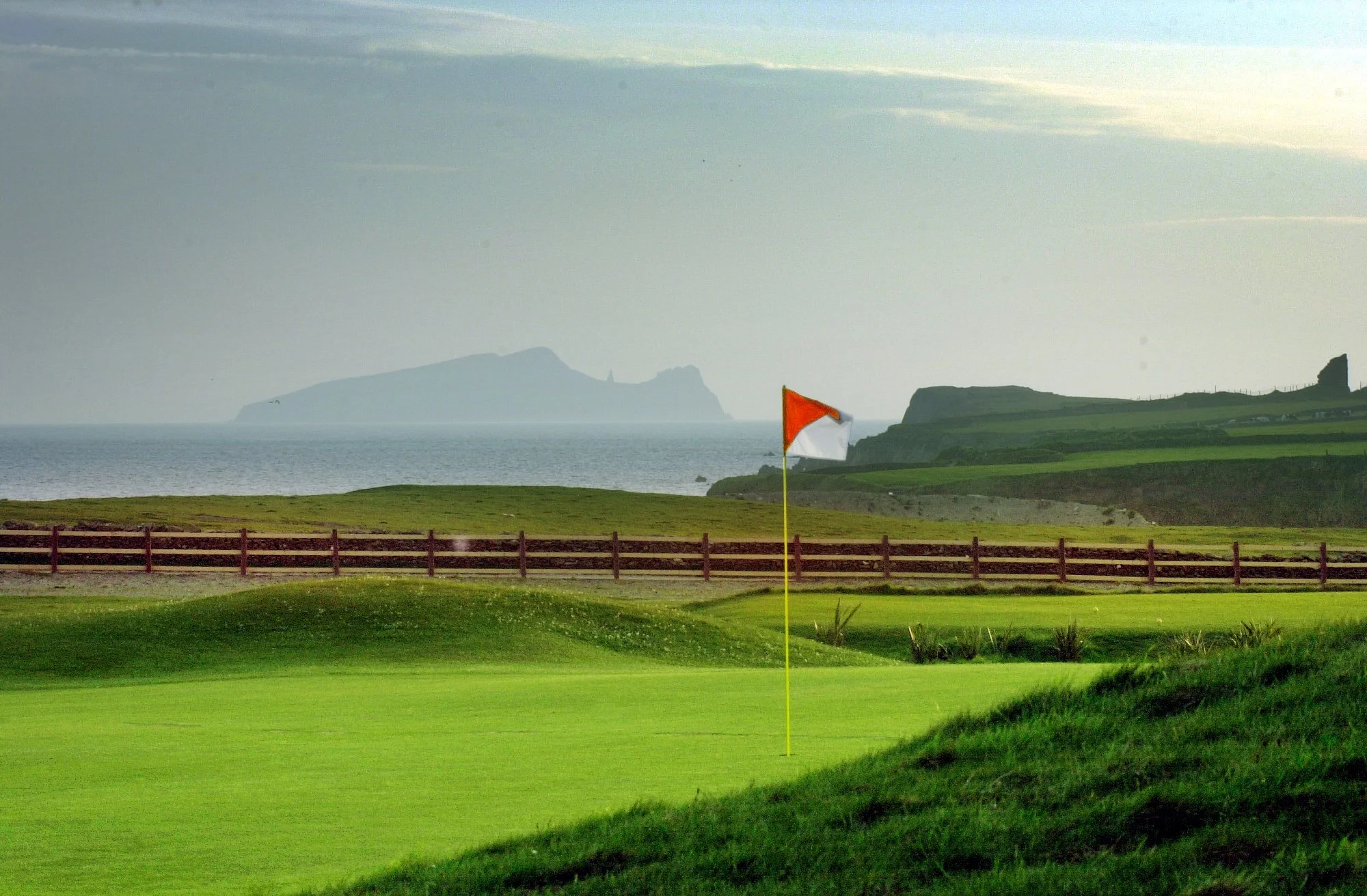 A golf course green with a flag near the hole, green grass, a wooden fence, and hills with cliffs in the background, along with an ocean and distant island.