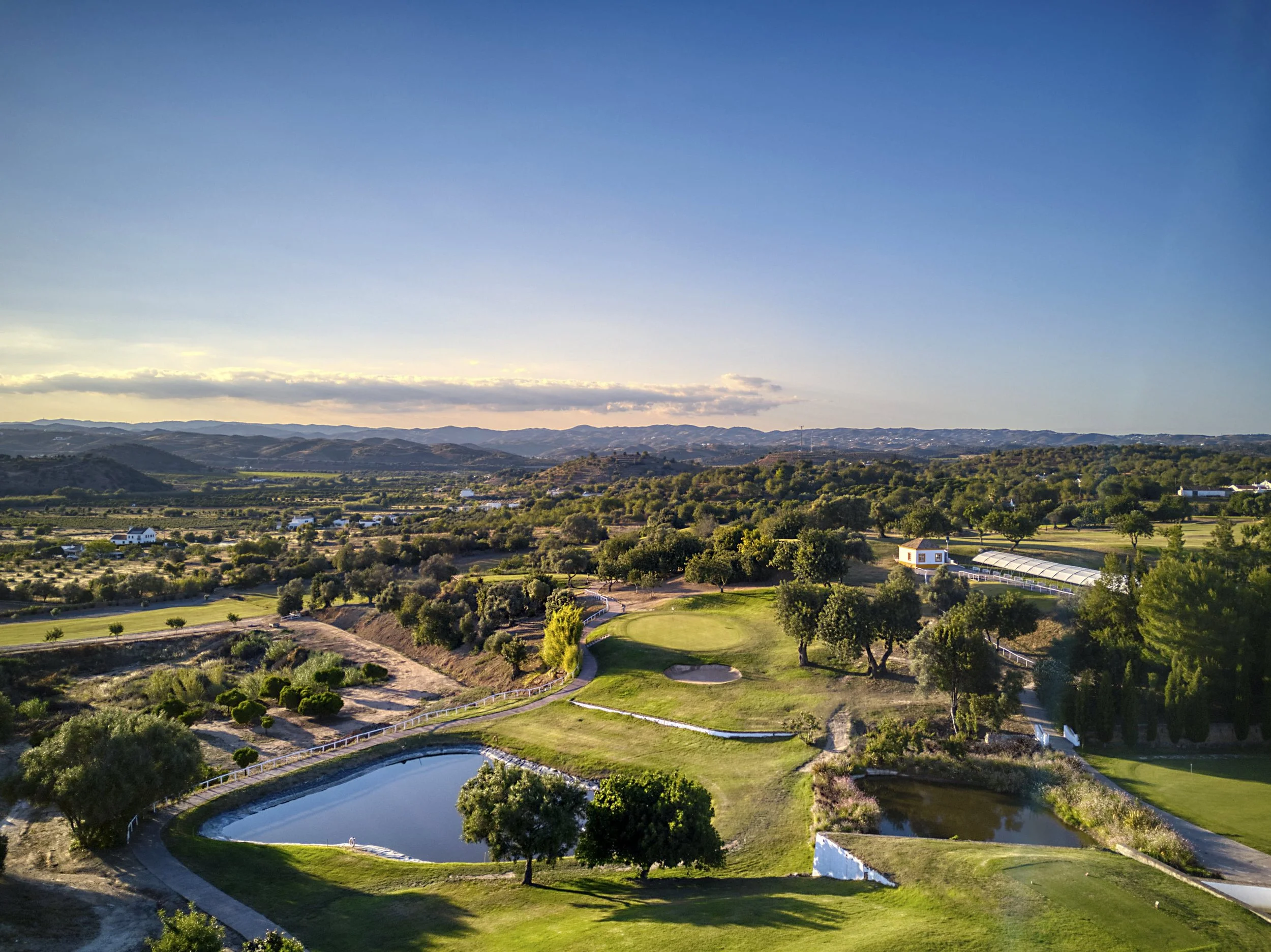 A scenic landscape of a golf course with ponds, trees, and rolling hills under a clear sky, taken during sunset.