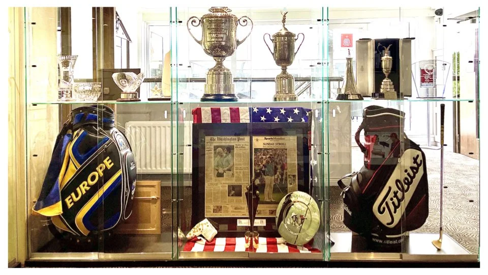 Display case with golf trophies, awards, and memorabilia, including golf bags from Europe and Titleist, framed newspapers, and a golf ball.