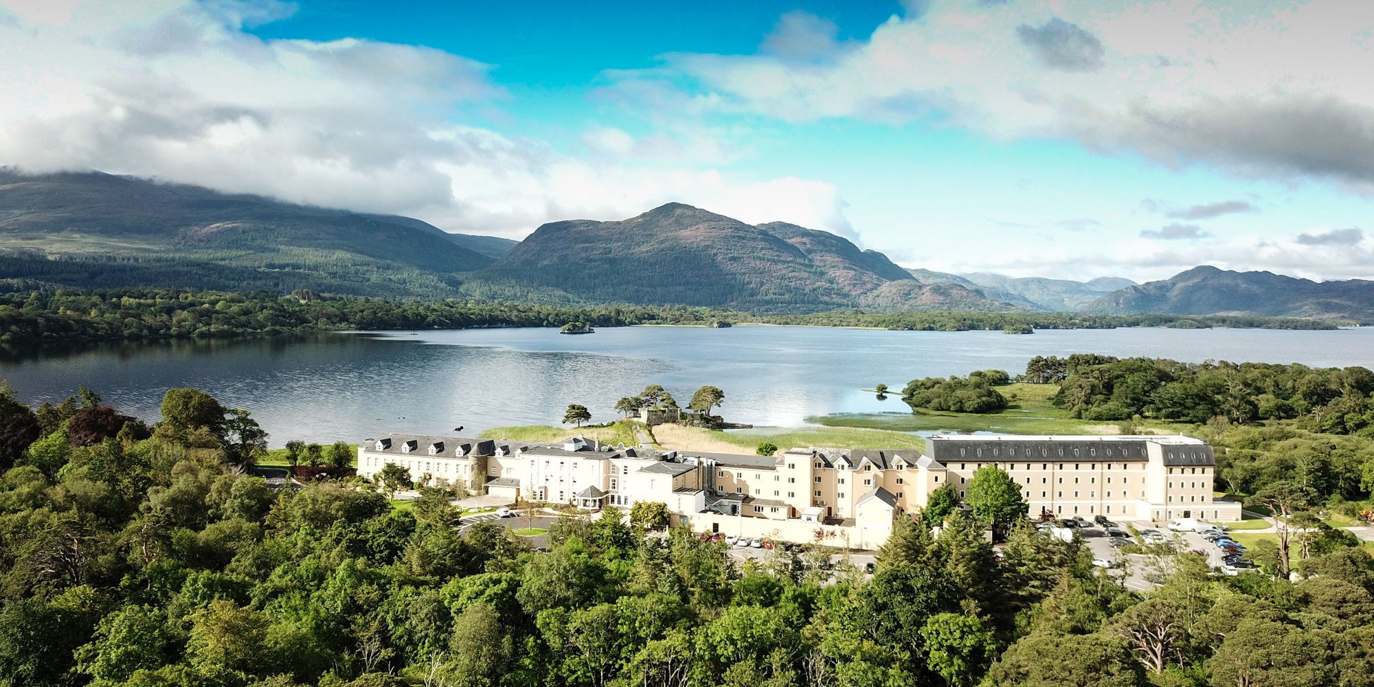 A scenic landscape featuring a large lake with surrounding greenery, mountains in the background, and a white building complex in the foreground.