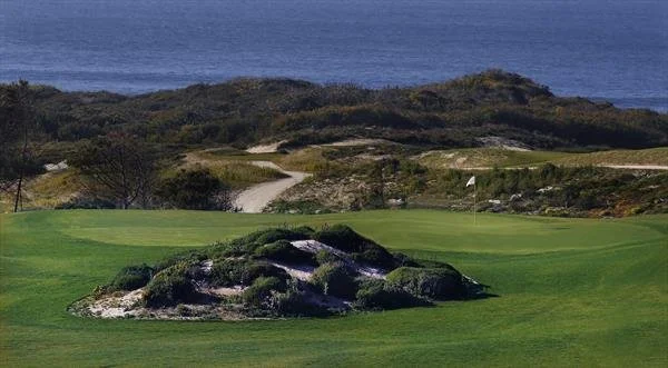 A golf course with a green closest to the camera, a small rocky island on the green, and a flag in the distance with the ocean in the background.