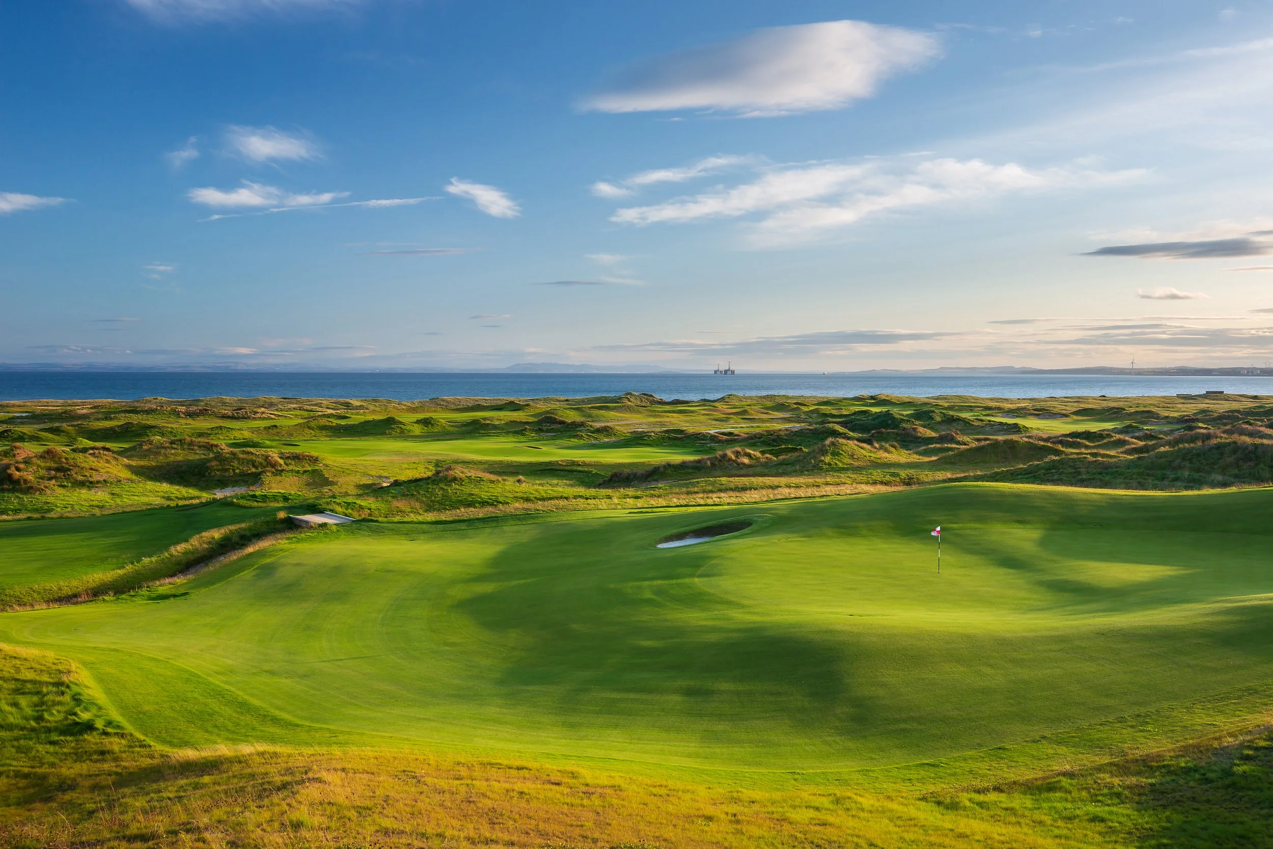 A scenic view of a golf course near the ocean on a clear day with blue skies and some clouds.