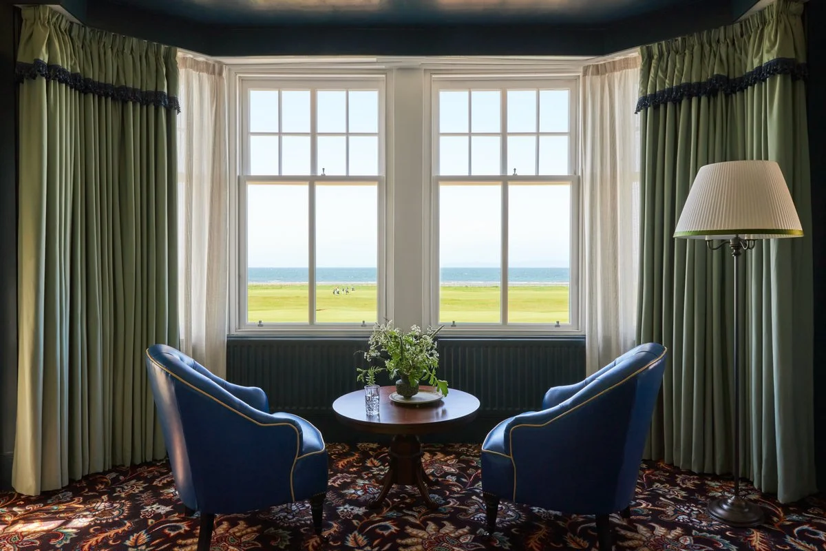 A cozy sitting area with two blue armchairs facing a large window showing a view of a golf course, grassy field, and ocean beyond, with curtains and a standing lamp nearby.