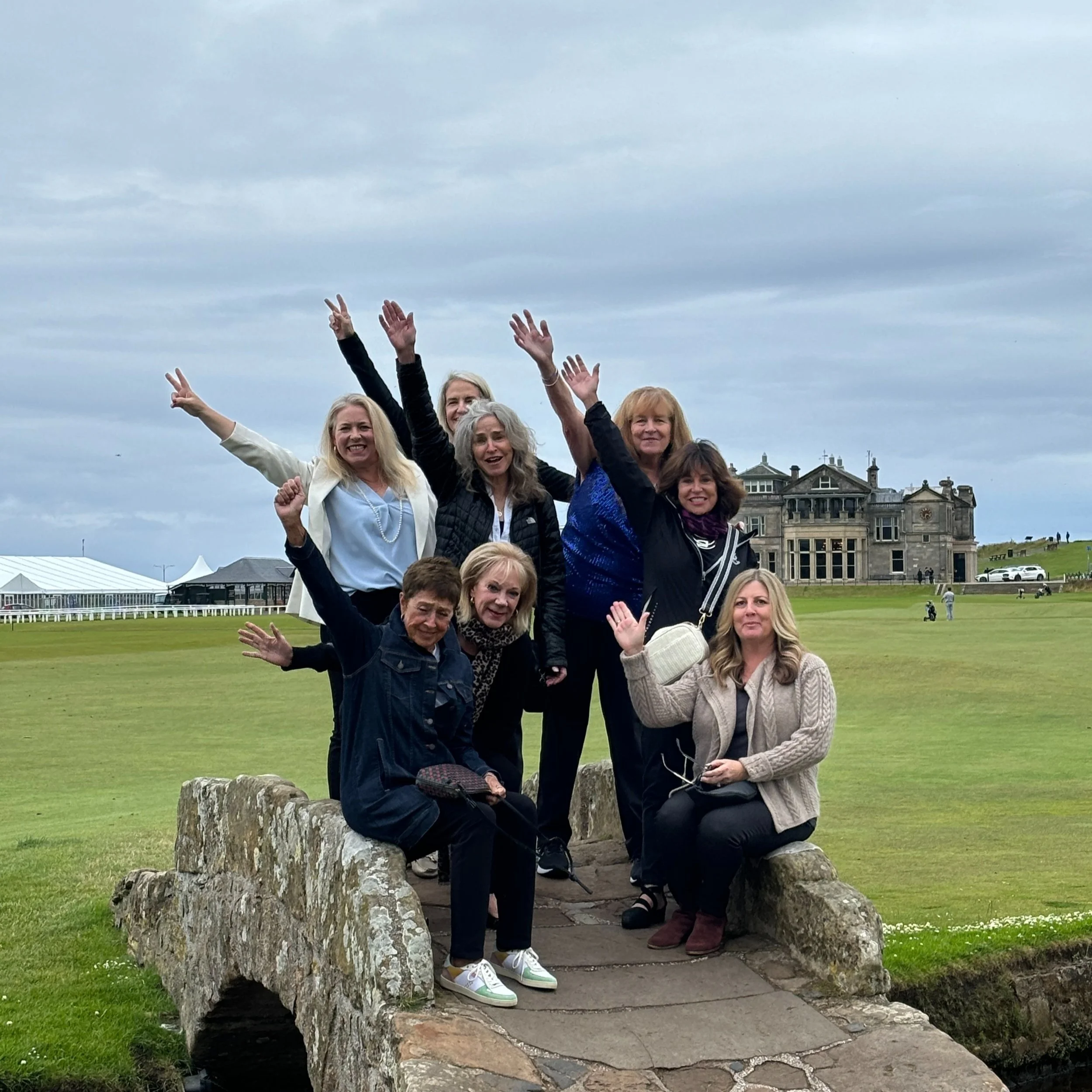 Group of women posing on a stone bridge at a golf course, with historic building in the background.