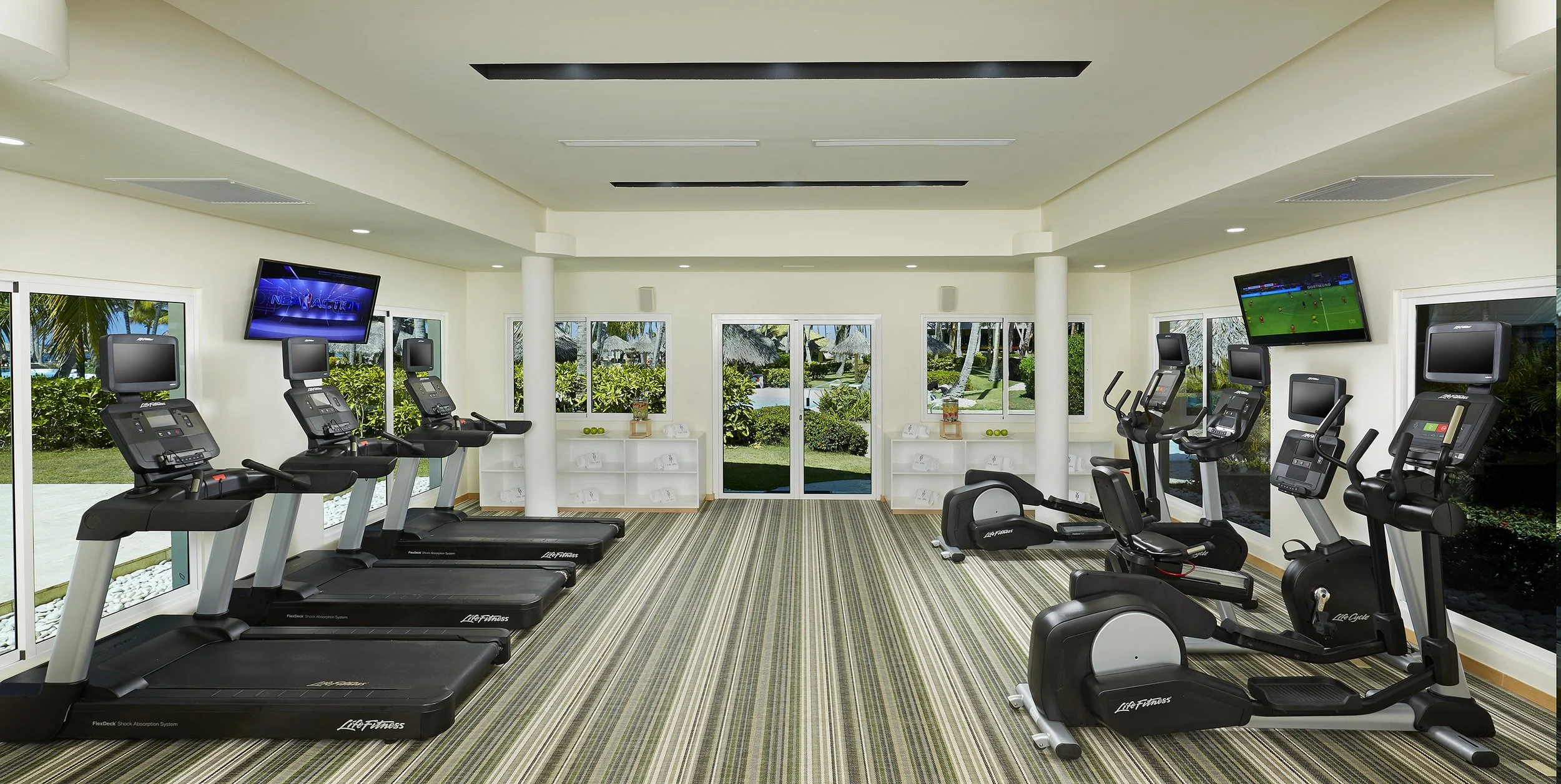 Indoor gym with treadmills, elliptical, and stationary bikes, large windows showing outdoor greenery, and ceiling-mounted TVs
