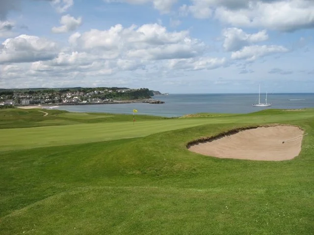 A view of a golf course with a green, a sand trap, and a flag, overlooking the ocean with sailboats and a coastal town in the distance under a partly cloudy sky.