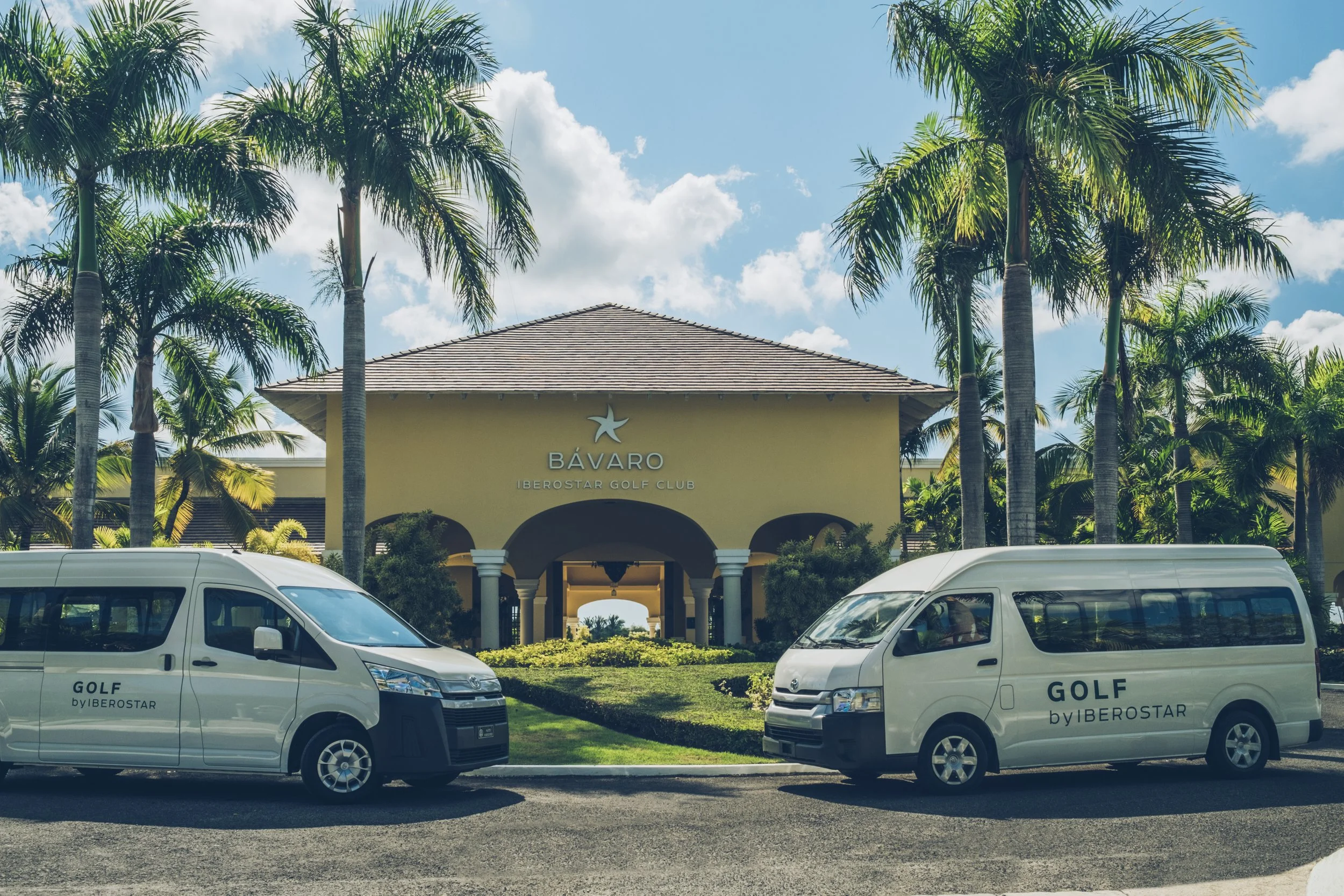 Entrance to Bávaro Iberostar Golf Club with two white golf shuttle vans parked in front, surrounded by palm trees under a partly cloudy sky.