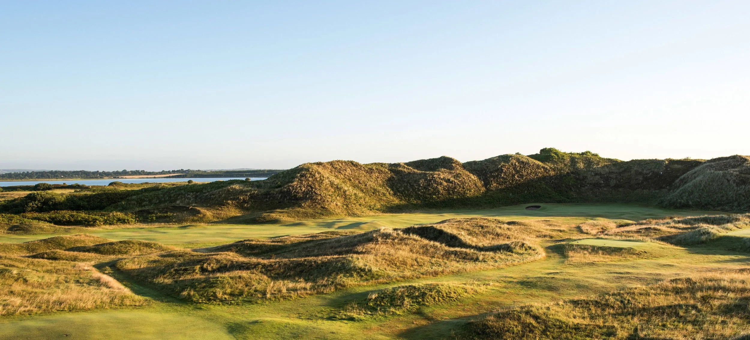 Sunlit view of a golf course with sand dunes and rolling grassy hills, with a body of water and trees in the distance under a clear blue sky.