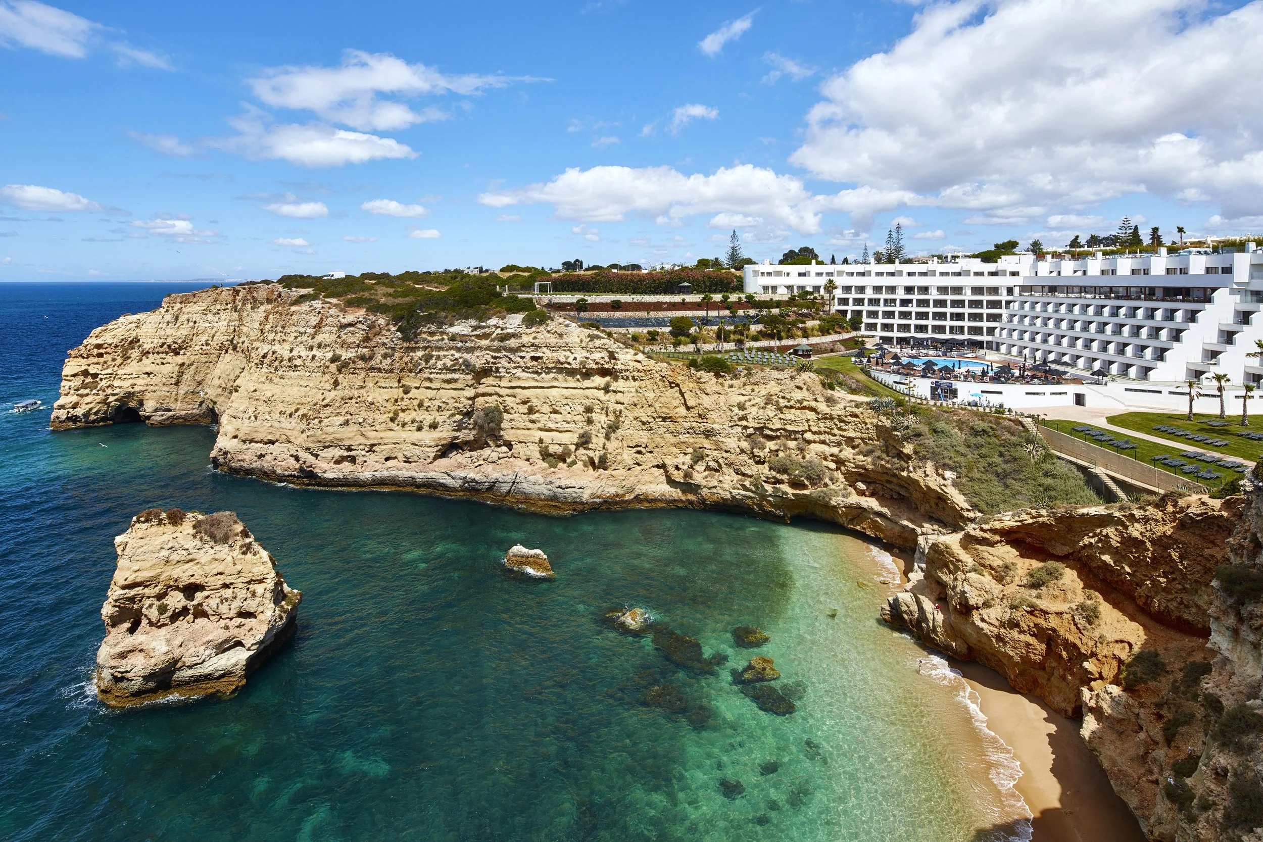 Coastal scene with a large white resort building, sandy beach, turquoise water, and rocky cliffs under a partly cloudy sky.