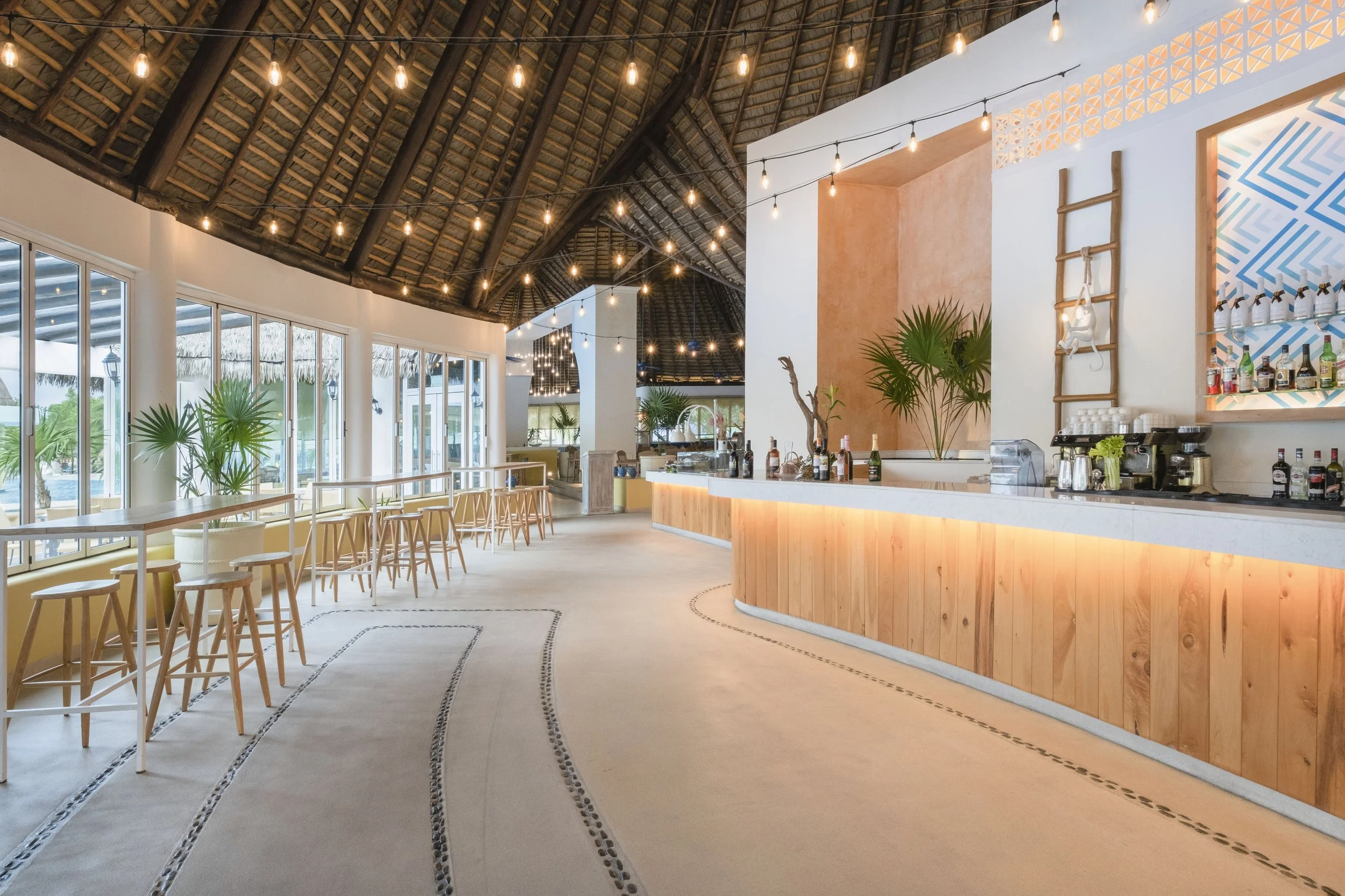 Interior of a tropical-themed restaurant or bar with wooden accents, string lights, and large windows showing greenery outside.