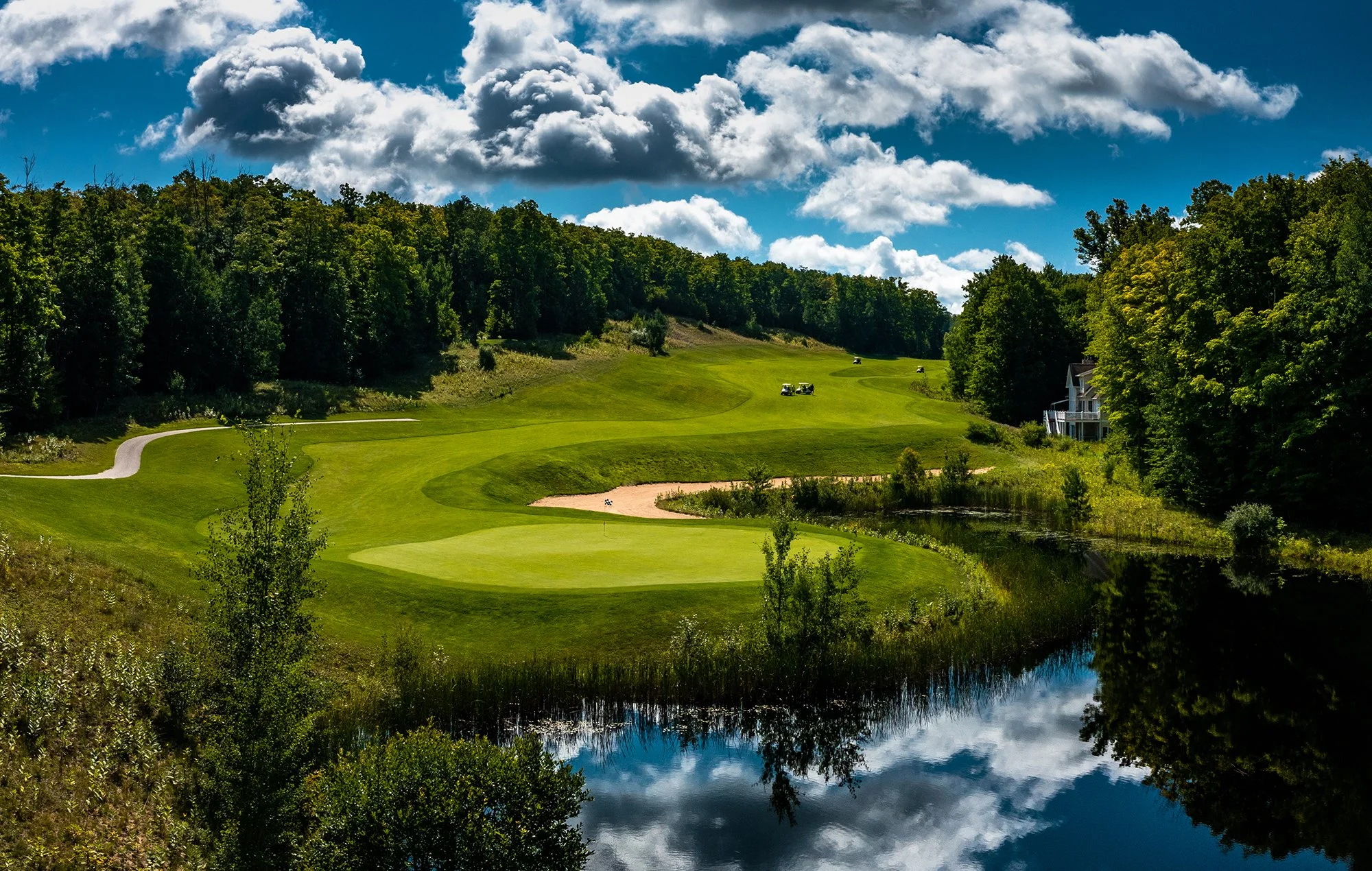 Panoramic view of a golf course with lush green fairways, sand traps, surrounded by dense trees, a bright blue sky with scattered clouds, and a calm water body reflecting the sky and trees.