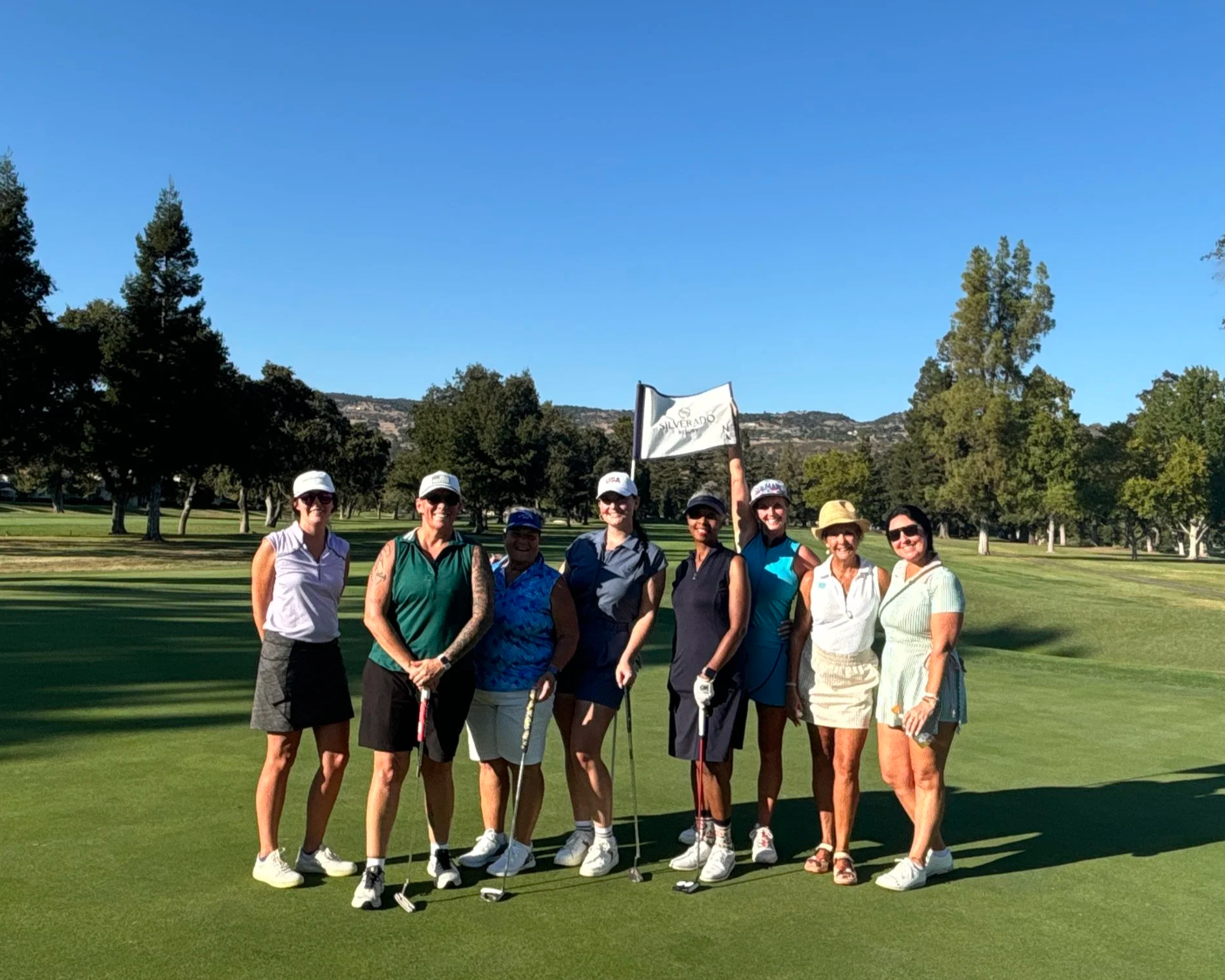 Group of nine women standing on a golf course, smiling, with a flag, trees, and mountains in the background.
