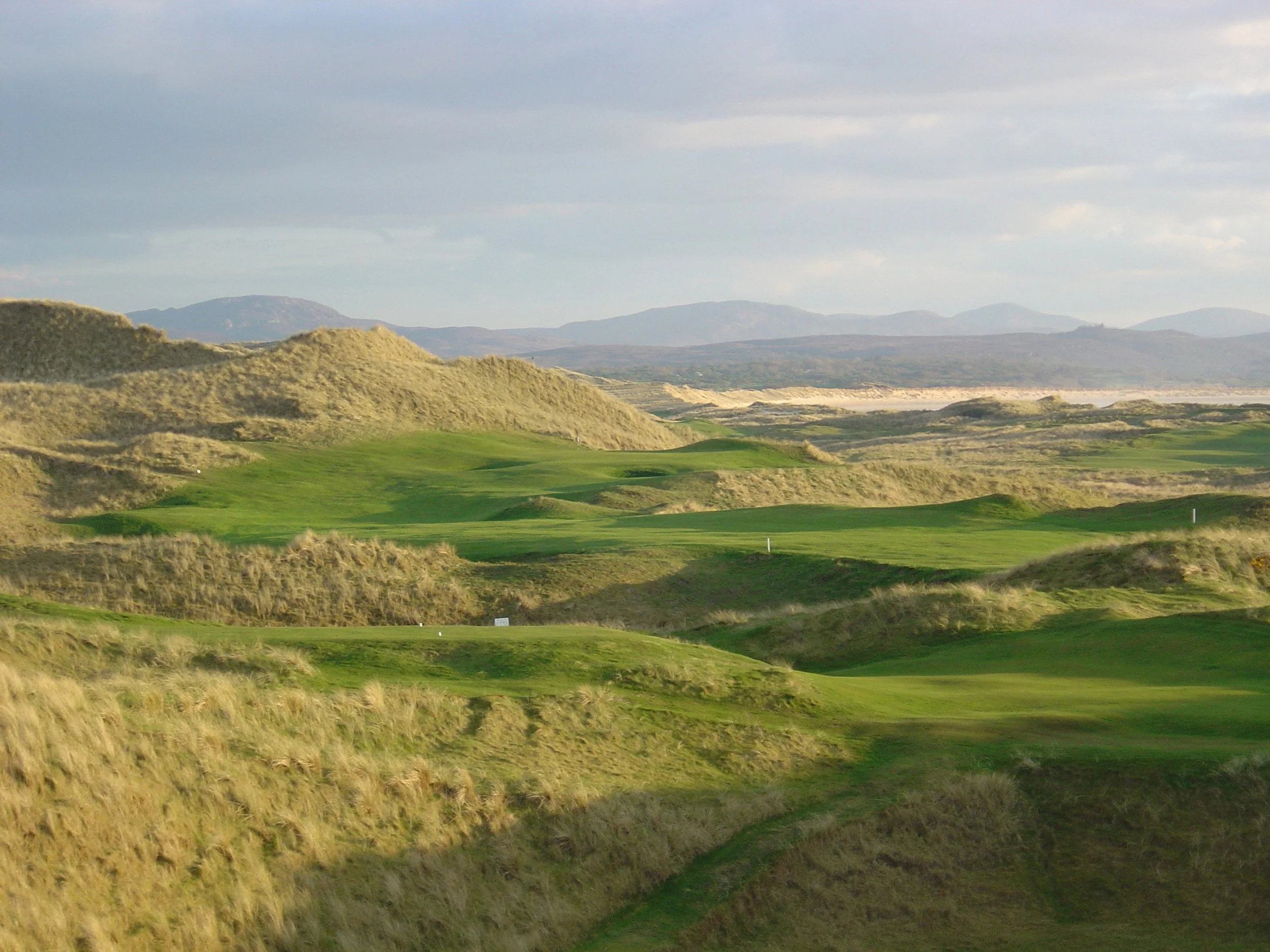 A scenic landscape of rolling green hills and sandy dunes under a partly cloudy sky.