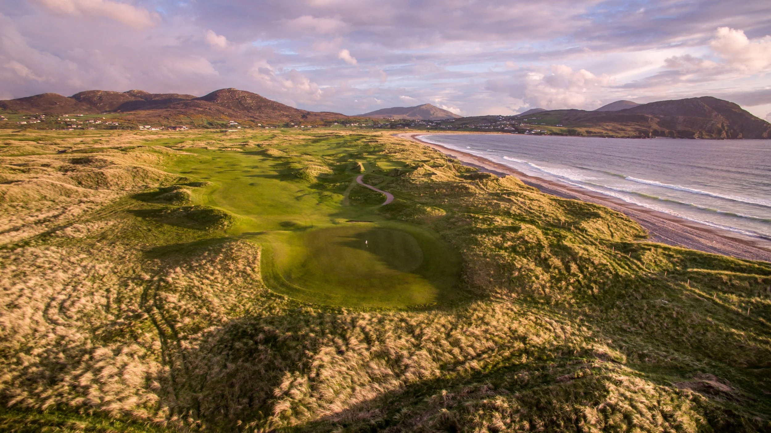 Aerial view of a coastal golf course with green fairways and sandy dunes, facing the ocean with waves and a beach, mountainous terrain in the background under a cloudy sky.