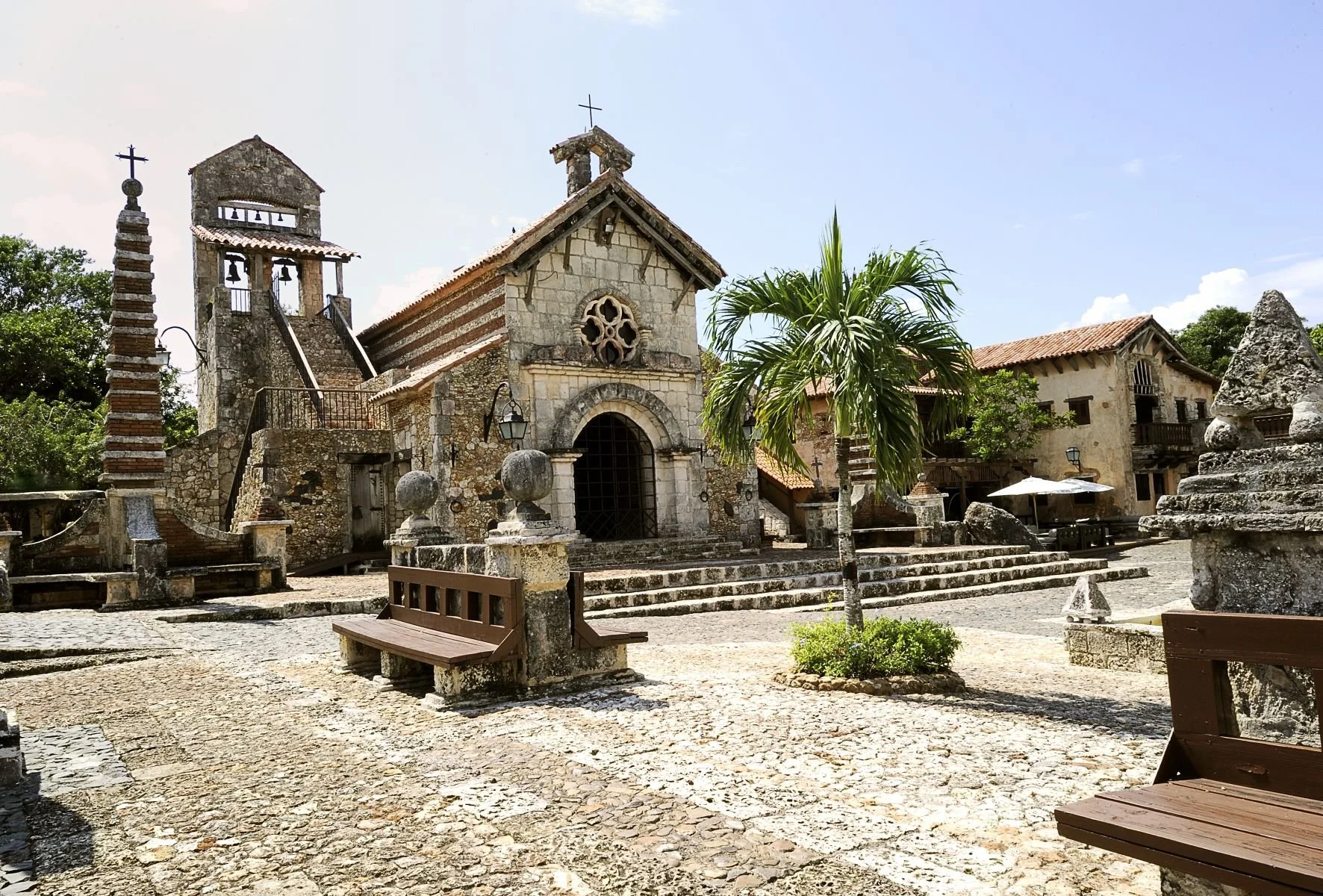 A historic stone church with a bell tower, surrounded by benches and a small palm tree in a sunny courtyard.