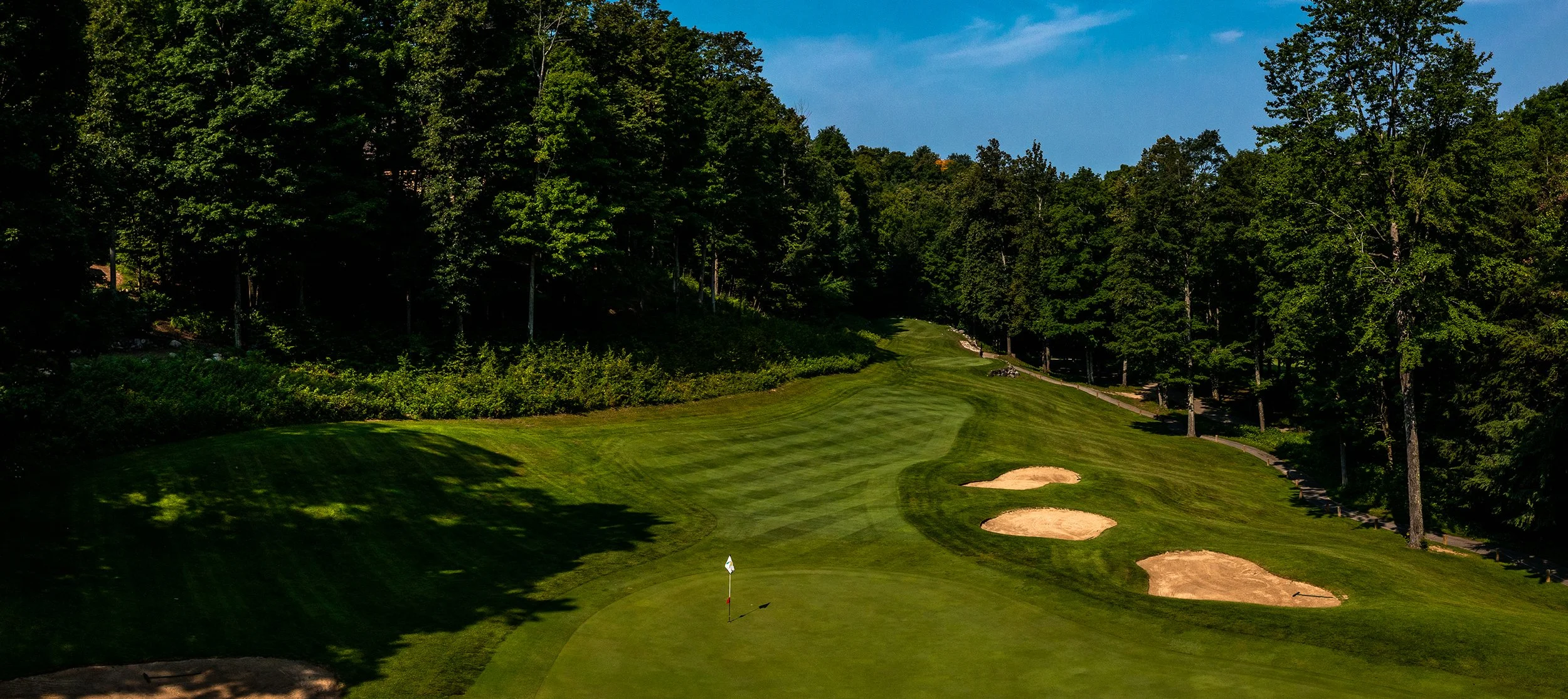 A golf course fairway with sand traps, surrounded by dense trees under a blue sky.