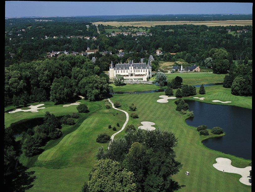 Aerial view of a golf course with sand traps, a pond, and lush green fairways, with a large mansion or estate in the background surrounded by trees.