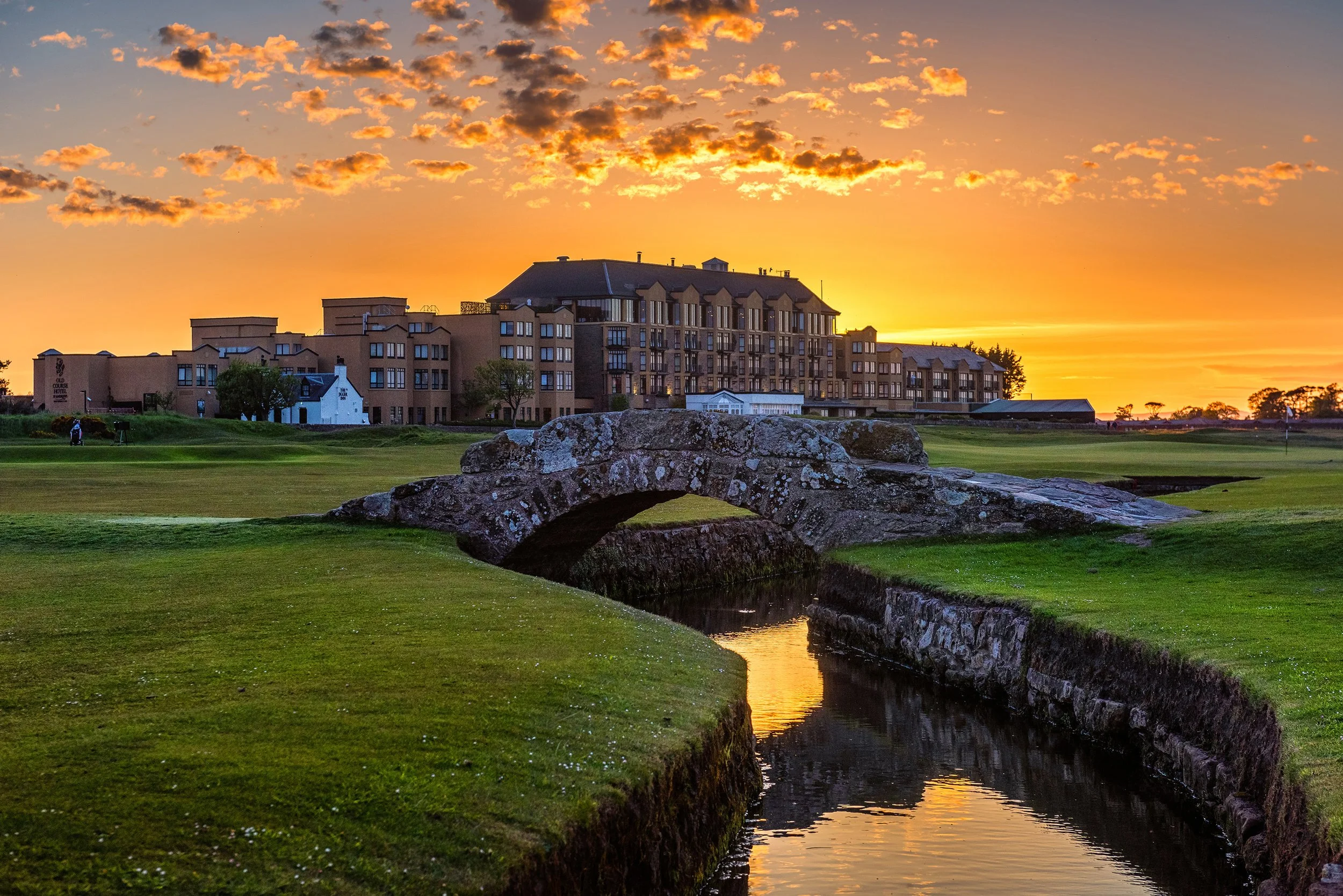 A brilliant golden sunset illuminates the historic Swilcan Bridge and the luxury Old Course Hotel overlooking the St Andrews Old Course fairways, a featured destination on our luxury escorted golf tours in Scotland.