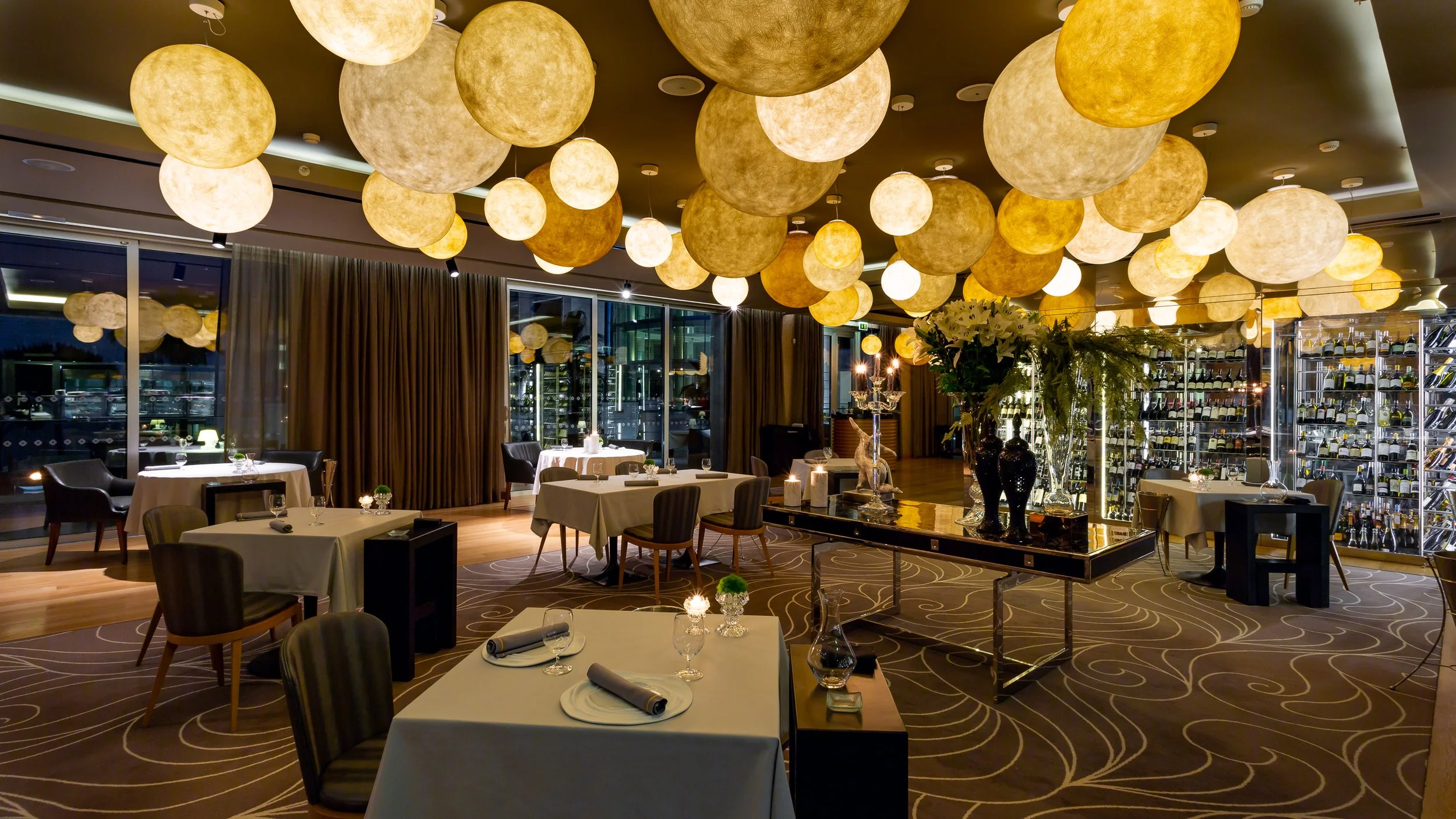 The elegant dining room of Gusto by Heinz Beck at the Conrad Algarve, featuring tables set for fine dining under a striking ceiling of warm, glowing spherical lights, with a glass wine cellar in the background.