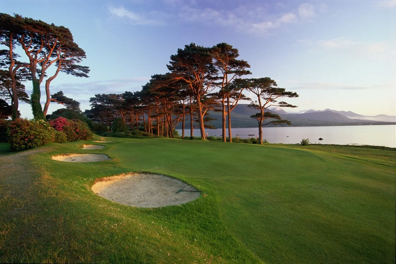 A scenic golf course near a lake at sunset, with trees and mountains in the background.