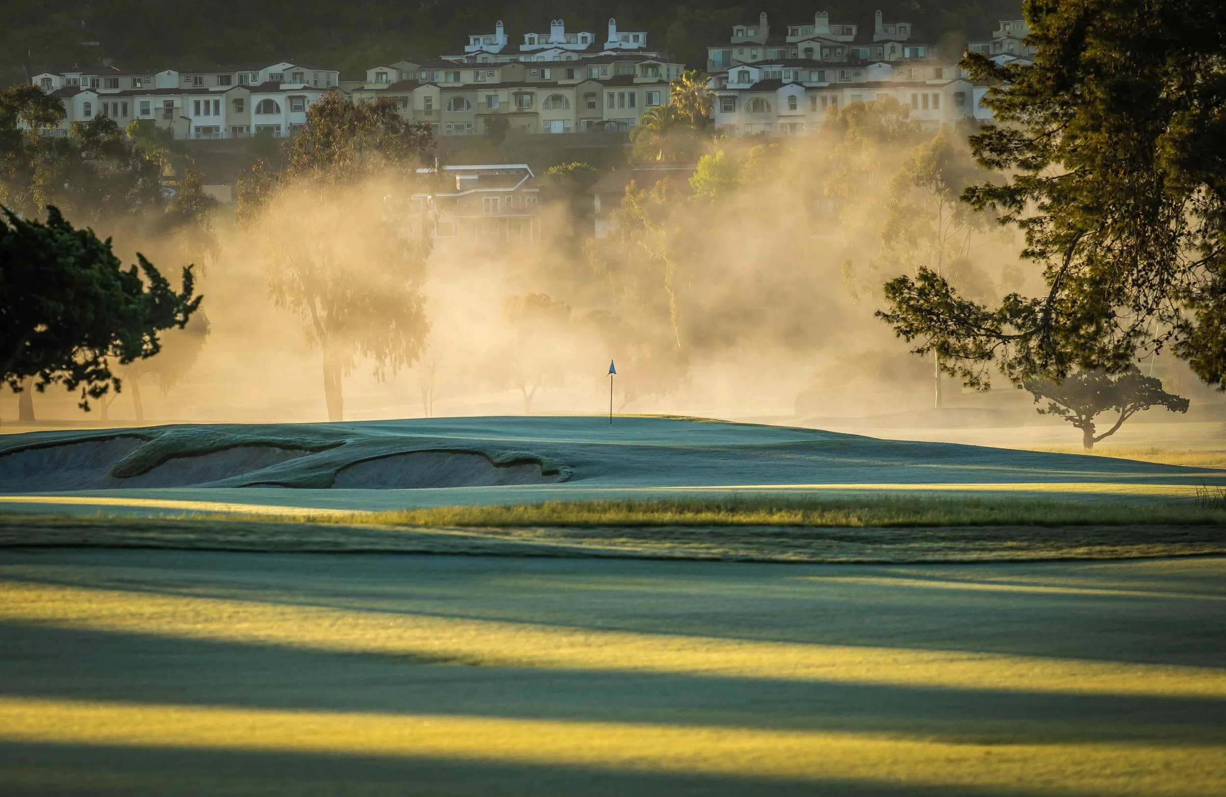 A golf course with a flag on the putting green, surrounded by trees and mist, with residential houses in the background during early morning or late evening.