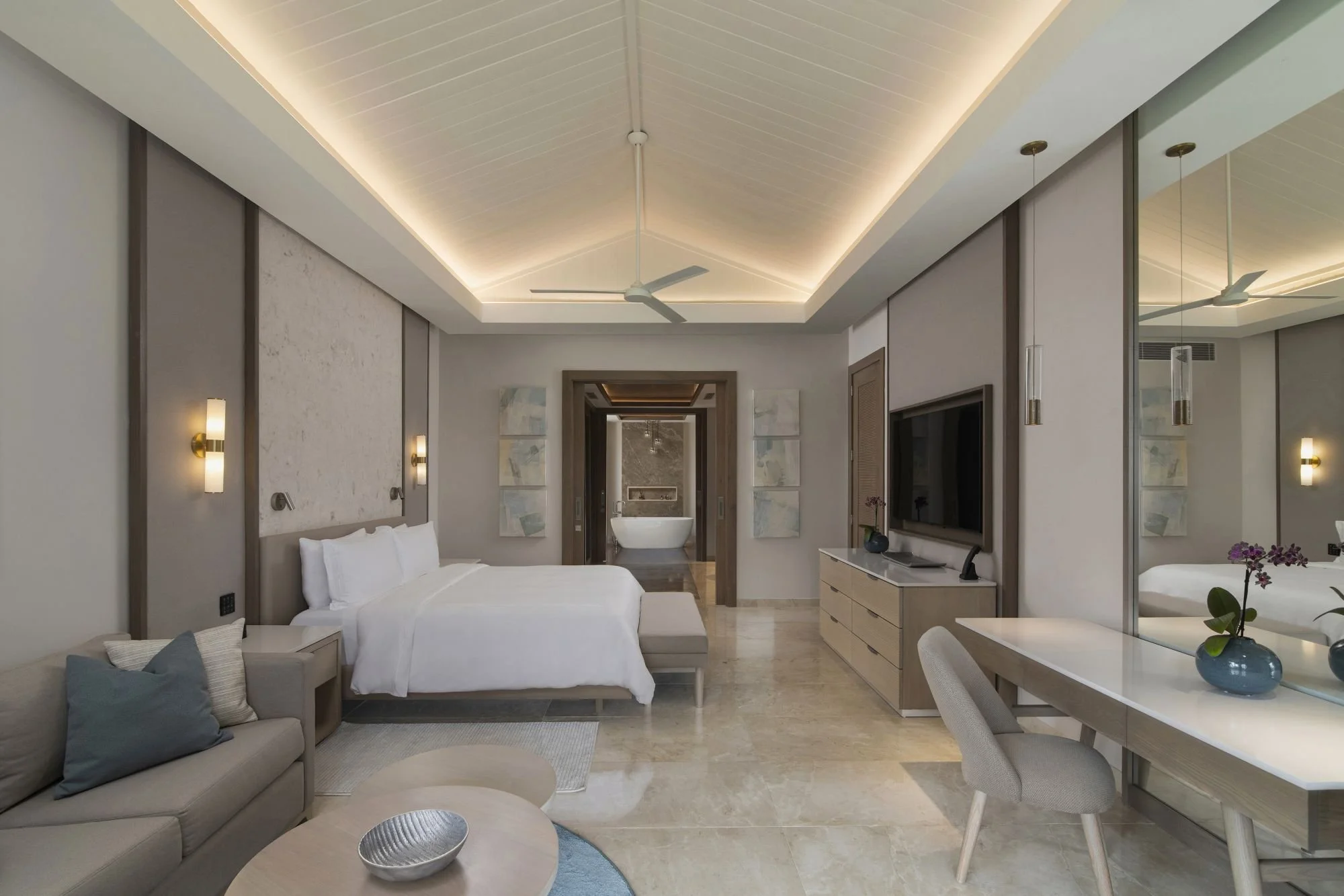 Modern hotel room with a bed, sitting area, TV, and a bathroom with a bathtub visible through an open doorway.